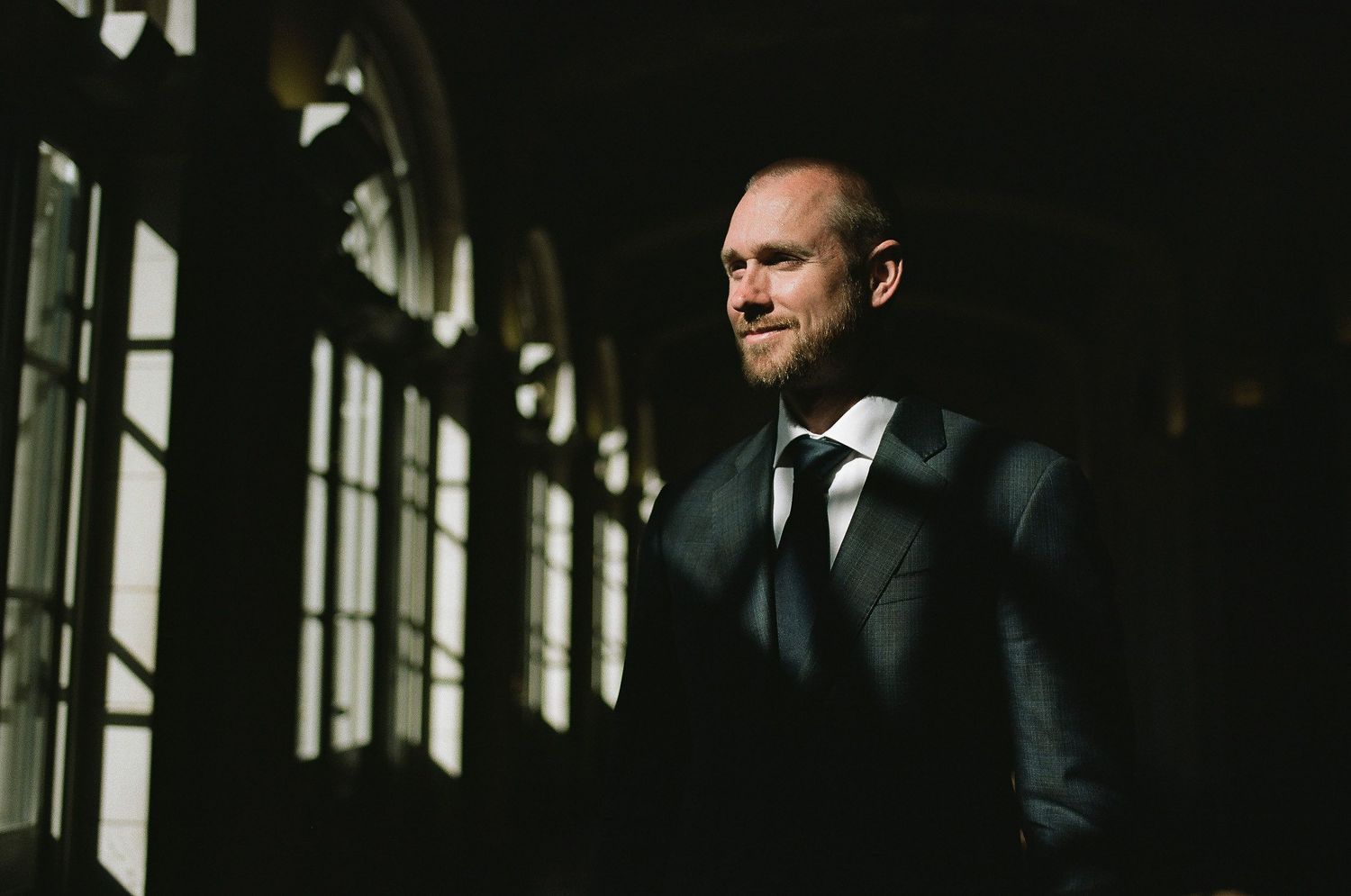 A person in a dark suit stands near large windows in moody, dramatic lighting creating a cinematic atmosphere.