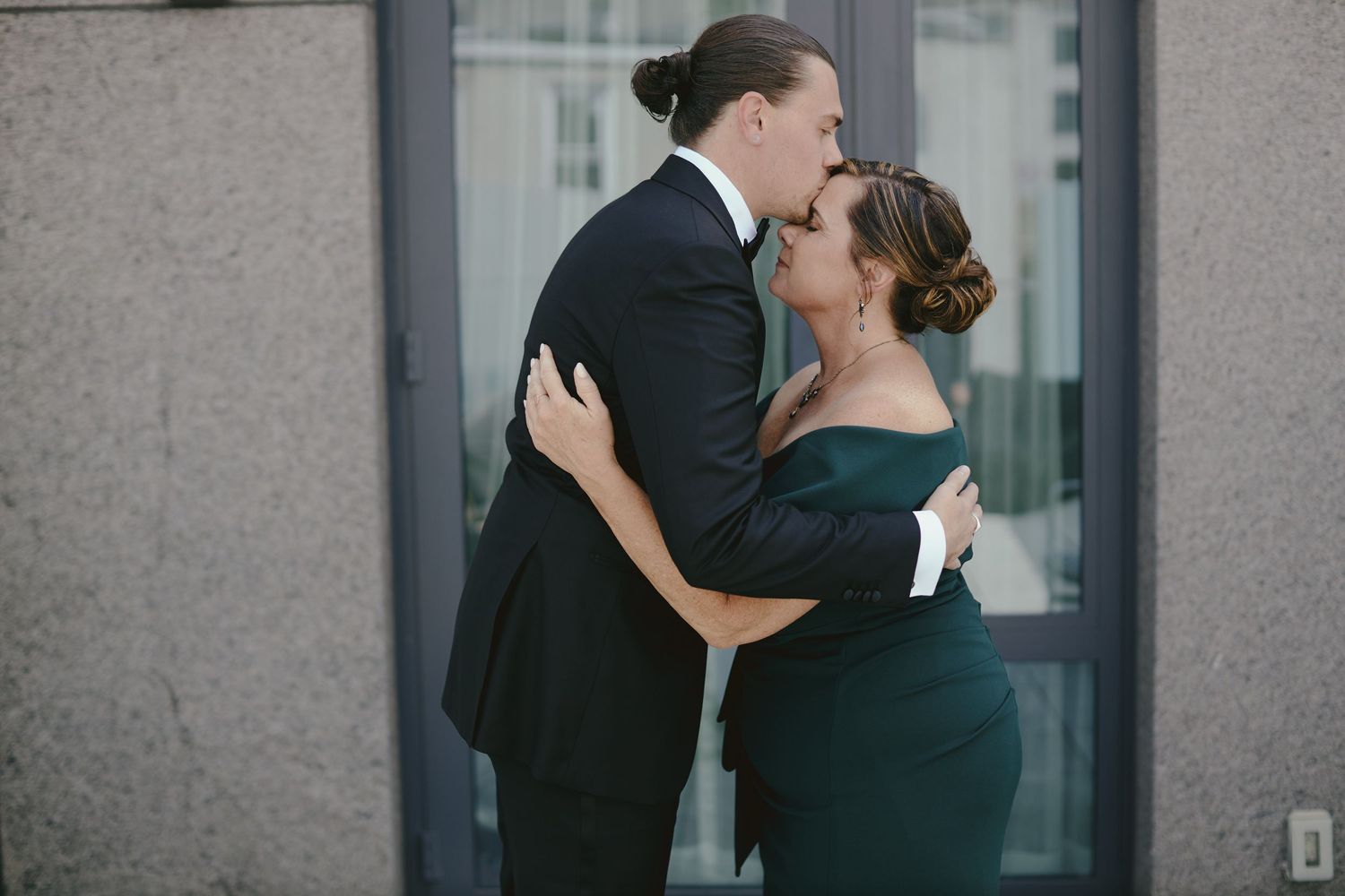 A romantic embrace between two people in formal black attire standing against a gray wall.