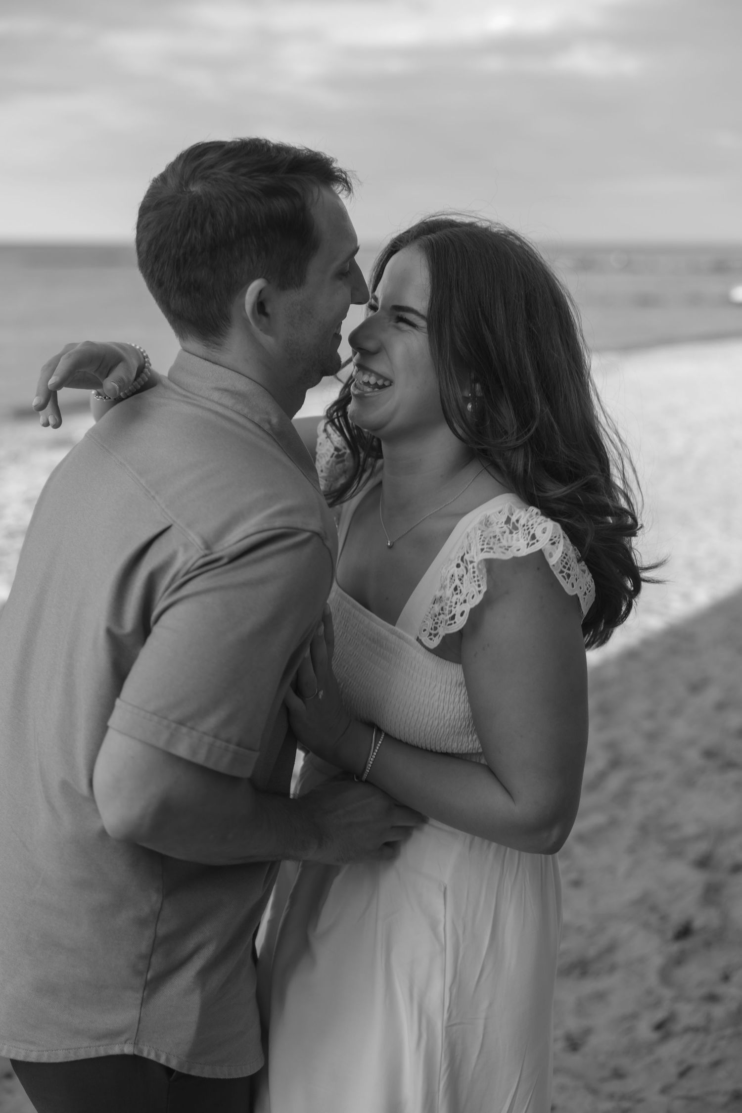 Black and white photo of a romantic couple embracing and smiling on a beach, with ocean waves in the background.