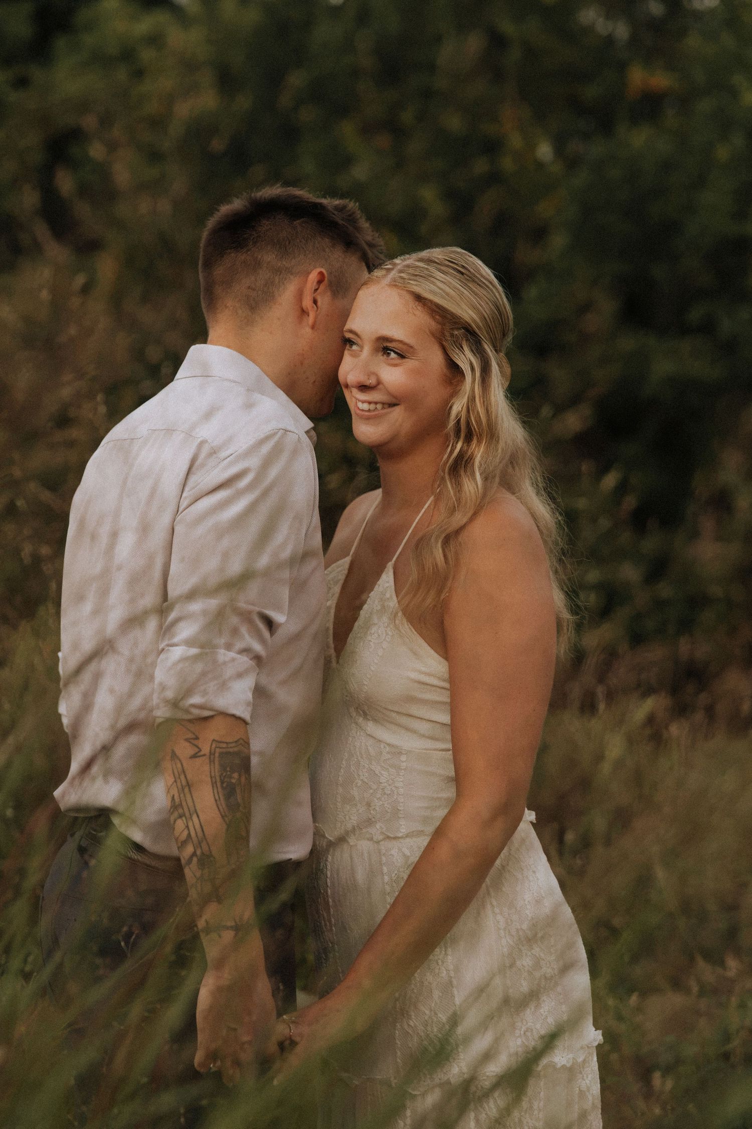 A smiling bride in a white lace dress holds hands with a tattooed groom in a field of green grass.