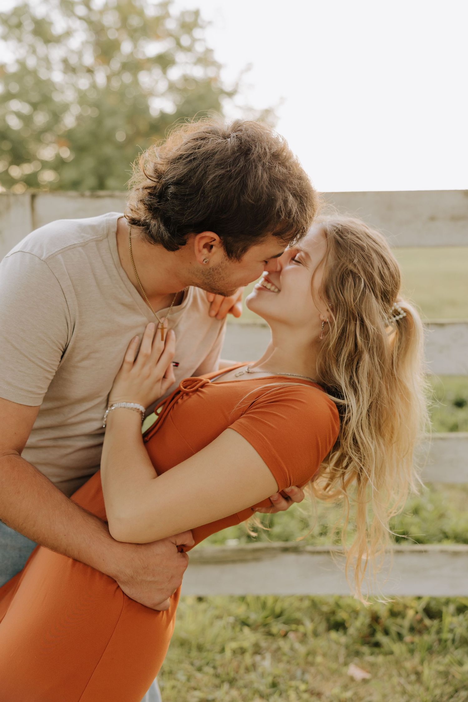 A romantic couple sharing intimate kisses in a sunny outdoor setting with green foliage in the background.