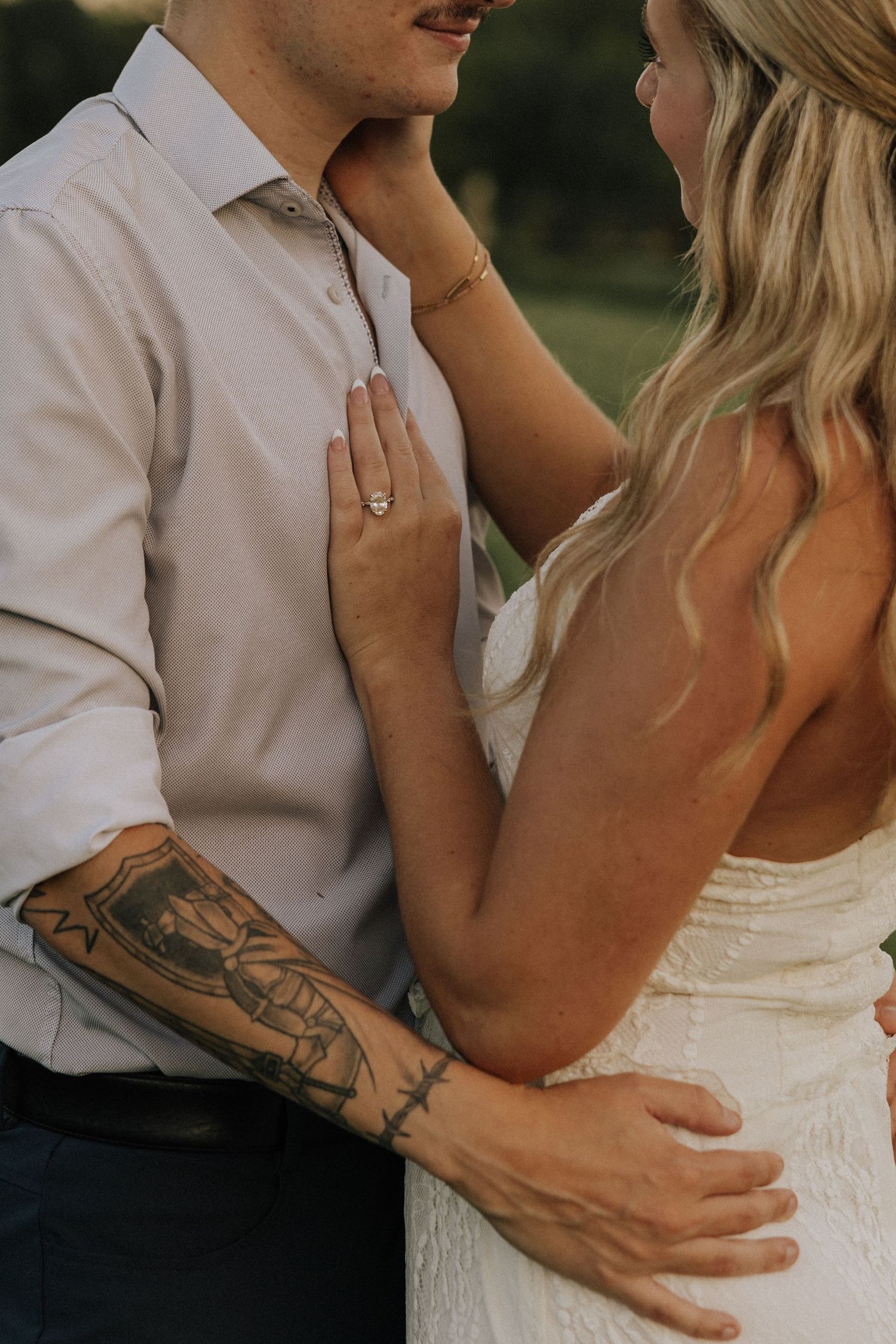 Bride with engagement ring and groom with tattoed arm embrace closely during outdoor wedding photo session.