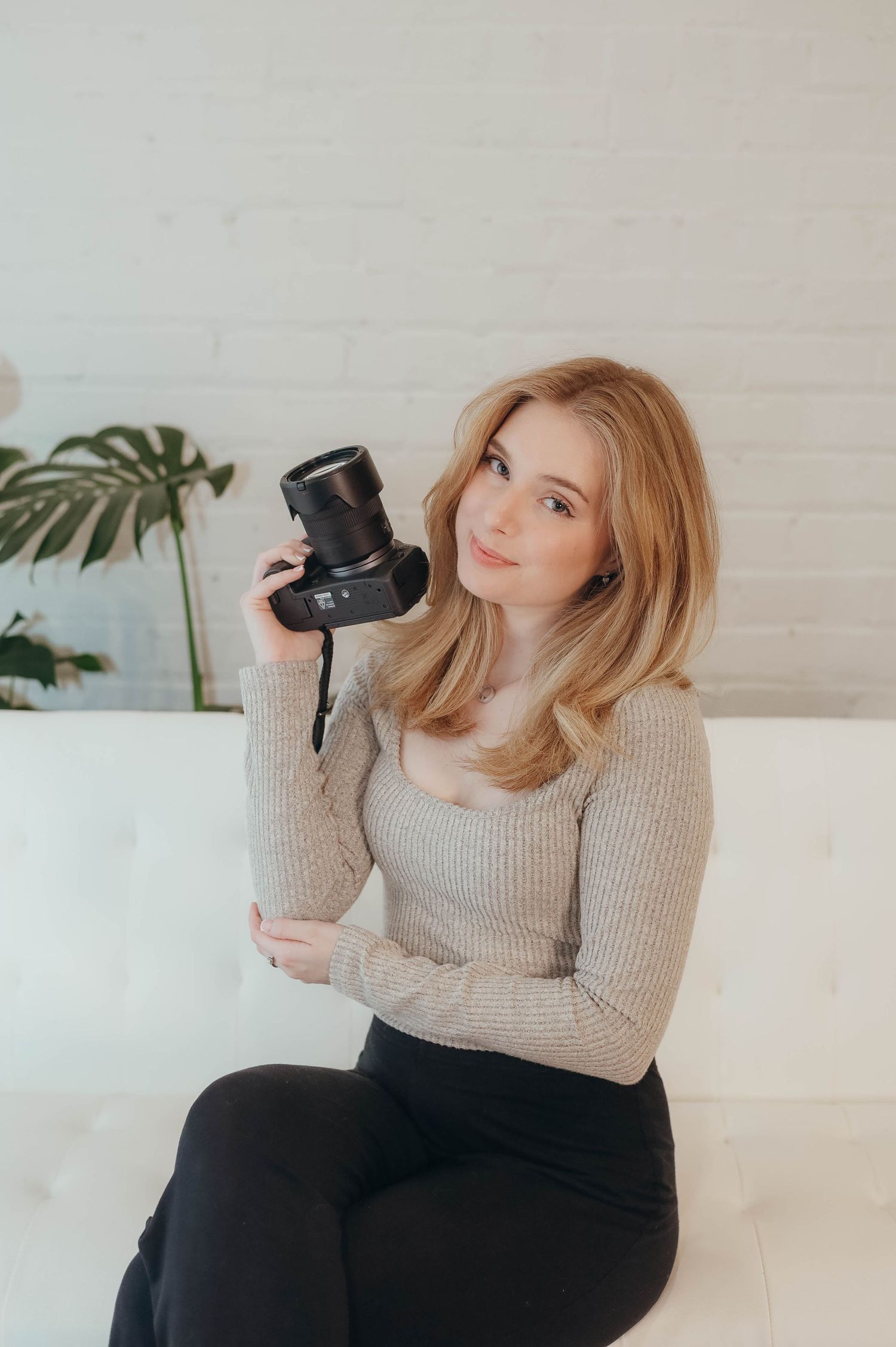 Photographer in a beige sweater and black pants poses with camera in minimalist white studio.