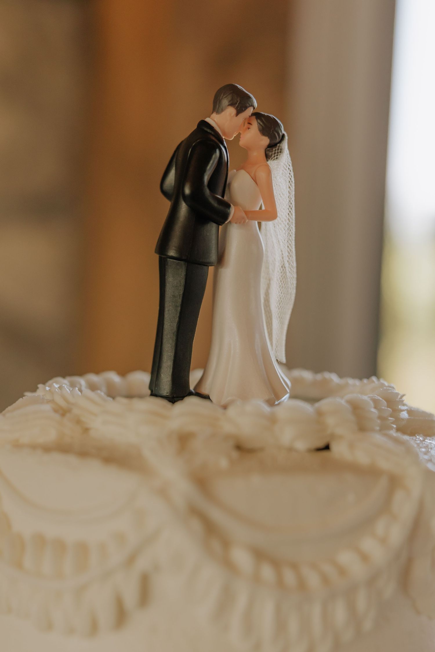 A traditional bride and groom wedding cake topper stands atop white frosted wedding cake with decorative piping details.