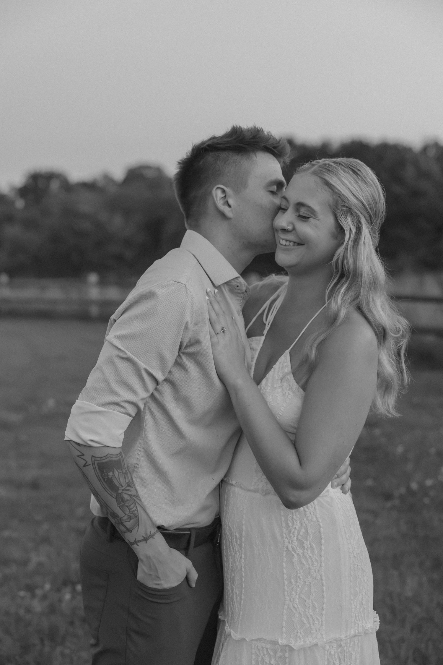 Black and white photo of a couple, man kissing woman on cheek outdoors, woman smiling in white dress.