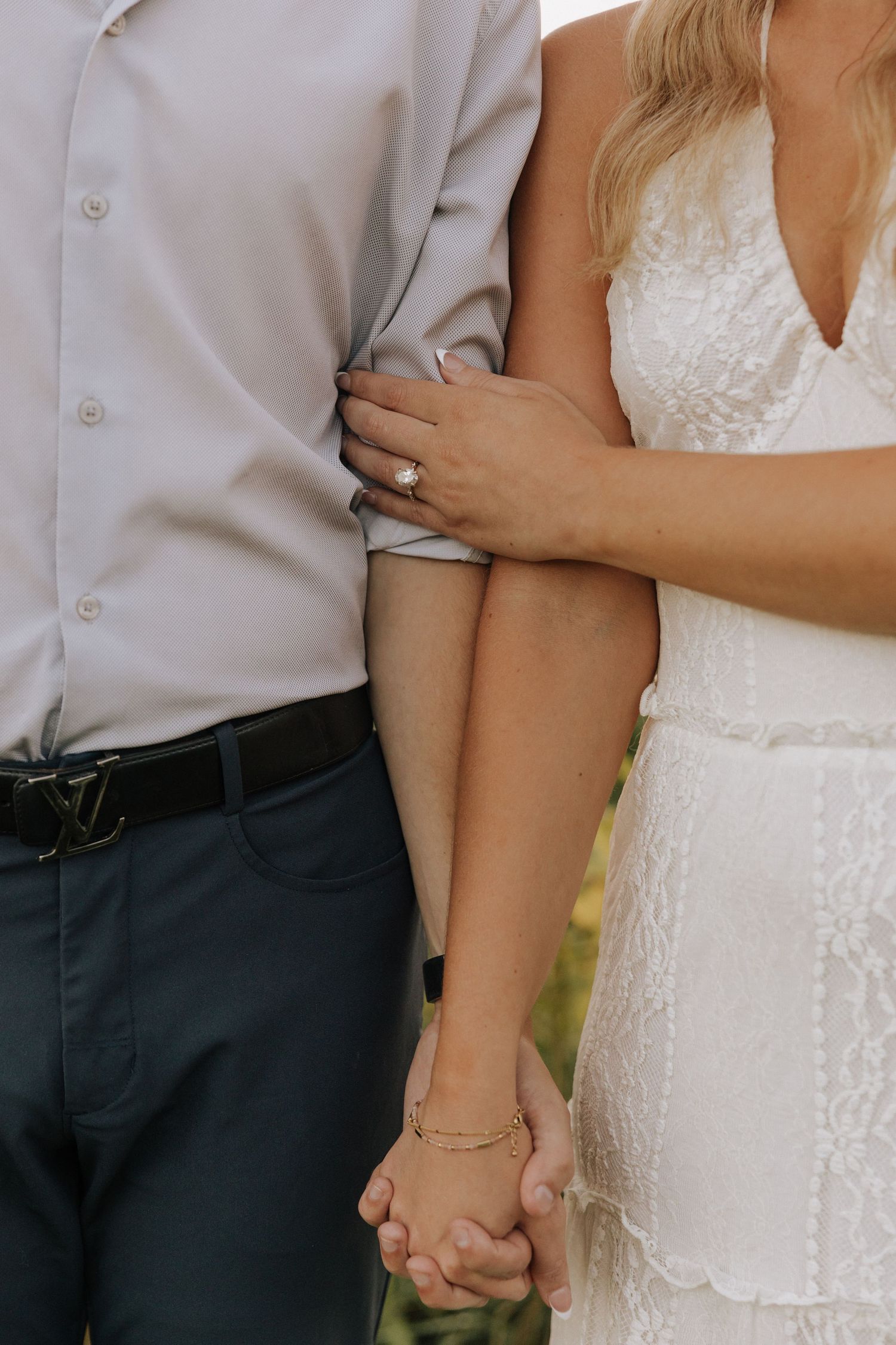 Couple holding hands with engagement ring visible, woman in white lace dress embracing man in gray shirt and dark pants.