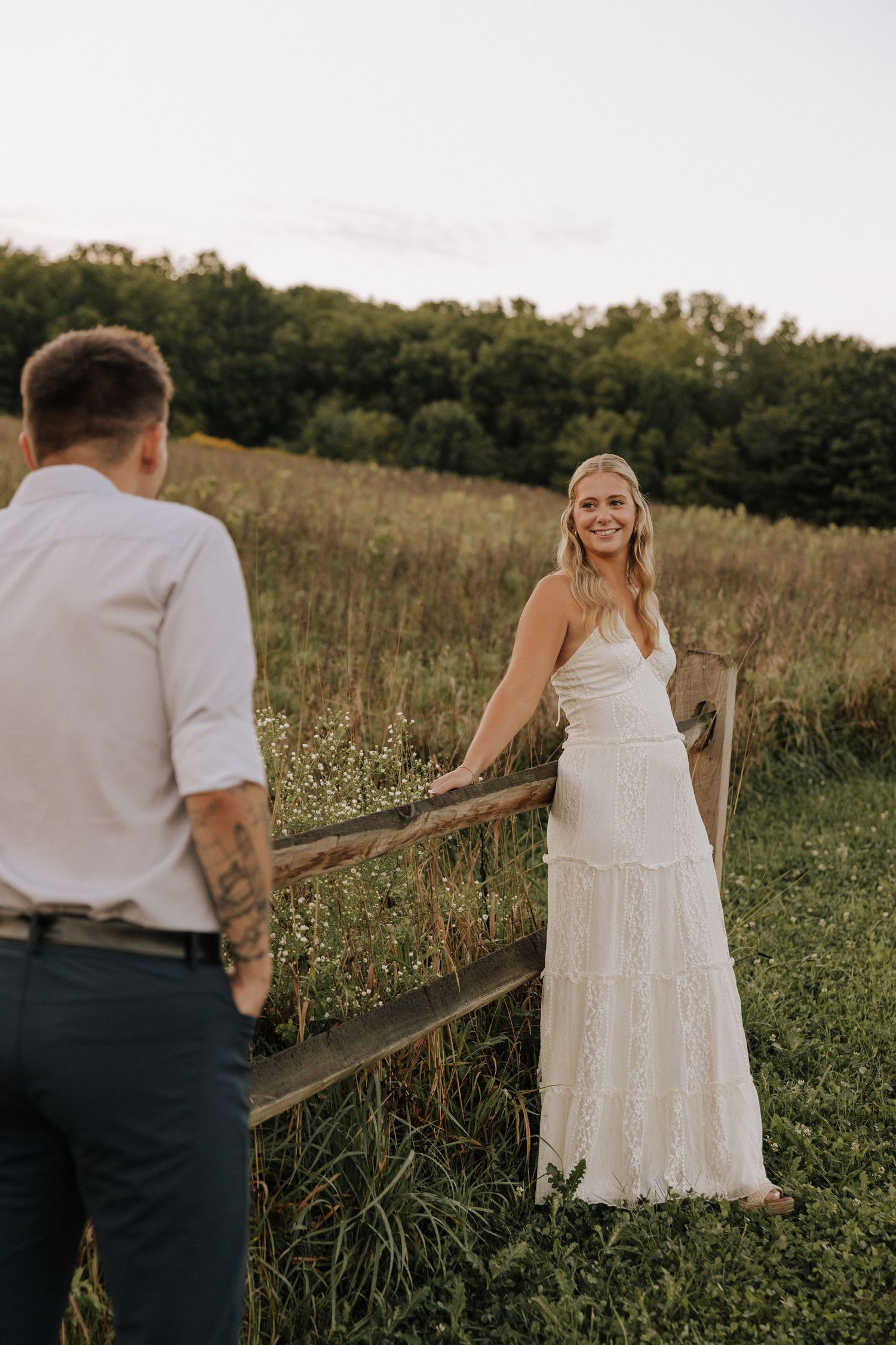 Bride in white lace gown smiles at groom by rustic fence in golden hour meadow.