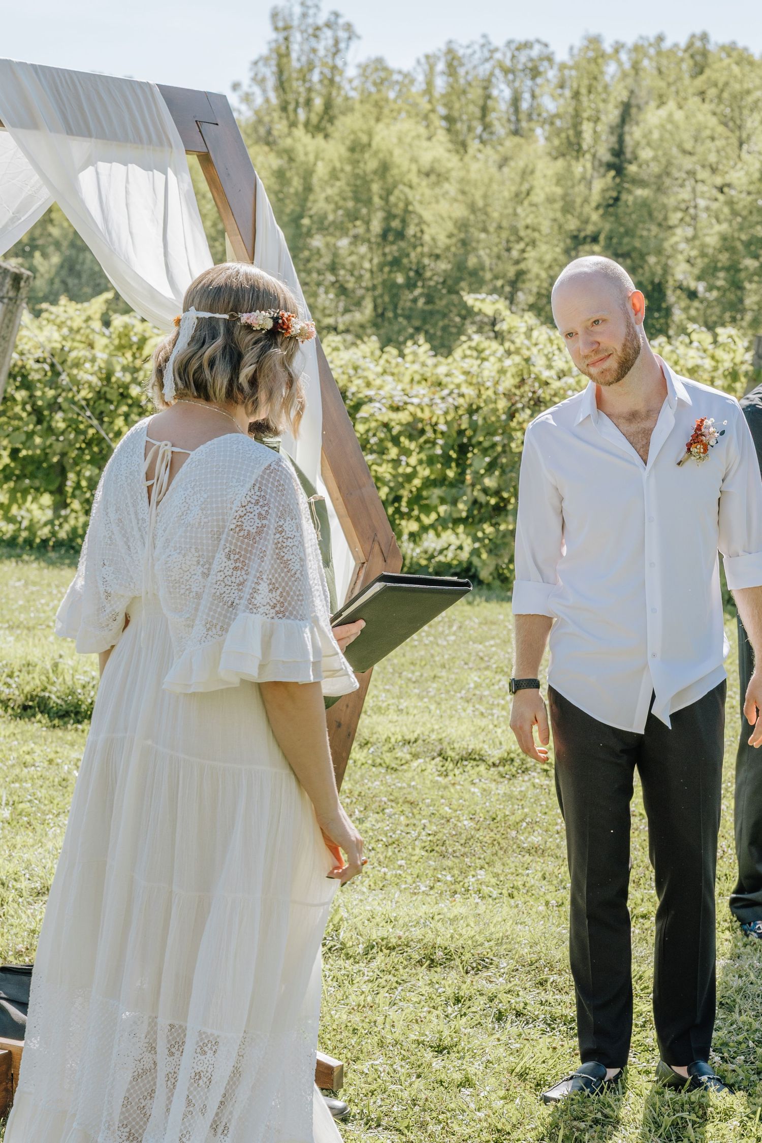 A couple exchanges vows during an outdoor summer wedding ceremony under a white fabric arch with greenery in background.