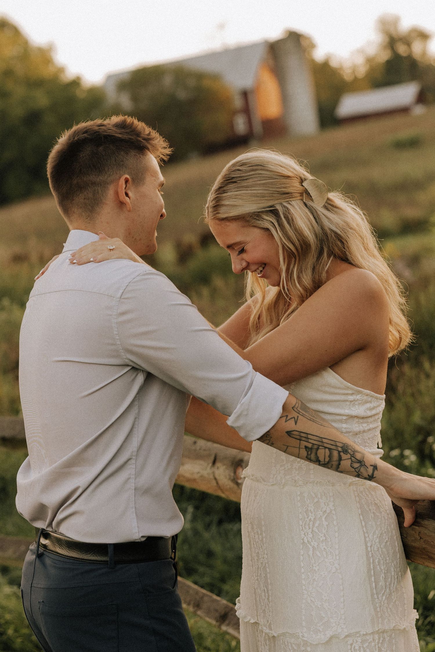 A couple shares a joyful moment outdoors at golden hour, with the woman in a white dress and visible tattoo near a wooden fence.