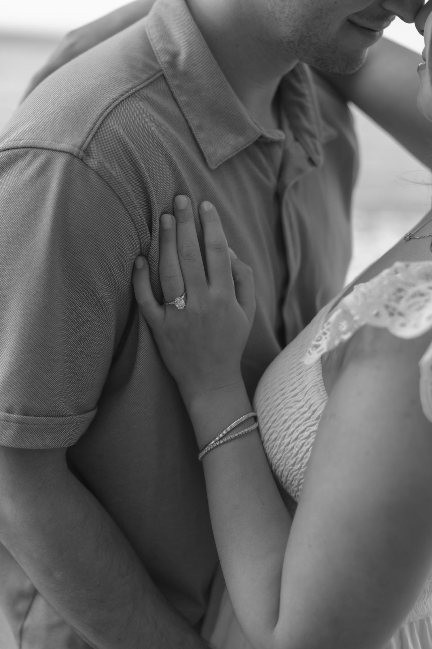 Black and white photo of couple embracing, woman's hand with engagement ring resting on man's chest.