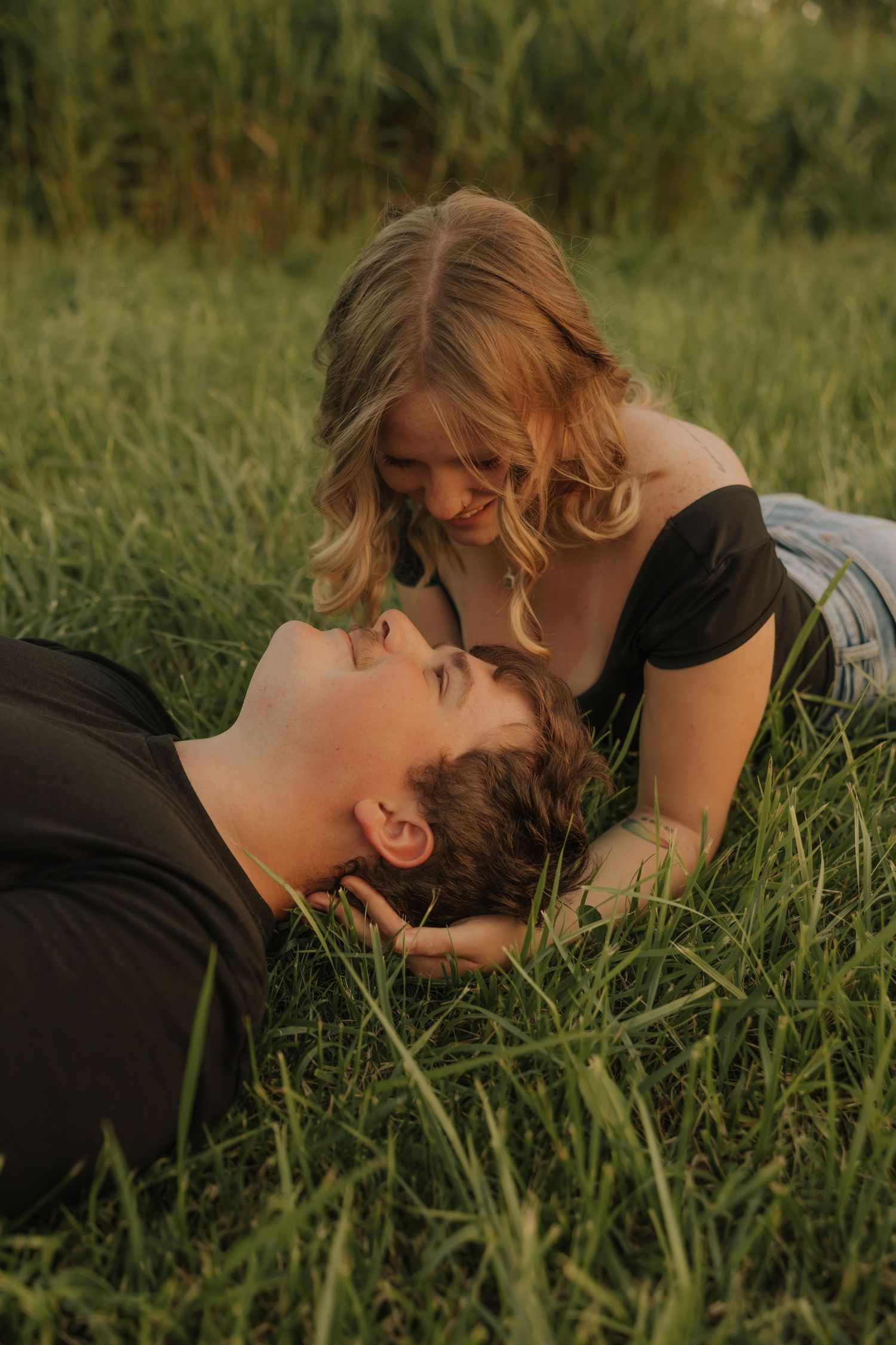 A couple shares a romantic moment lying in lush green grass, the woman smiling down at the man in warm golden light.