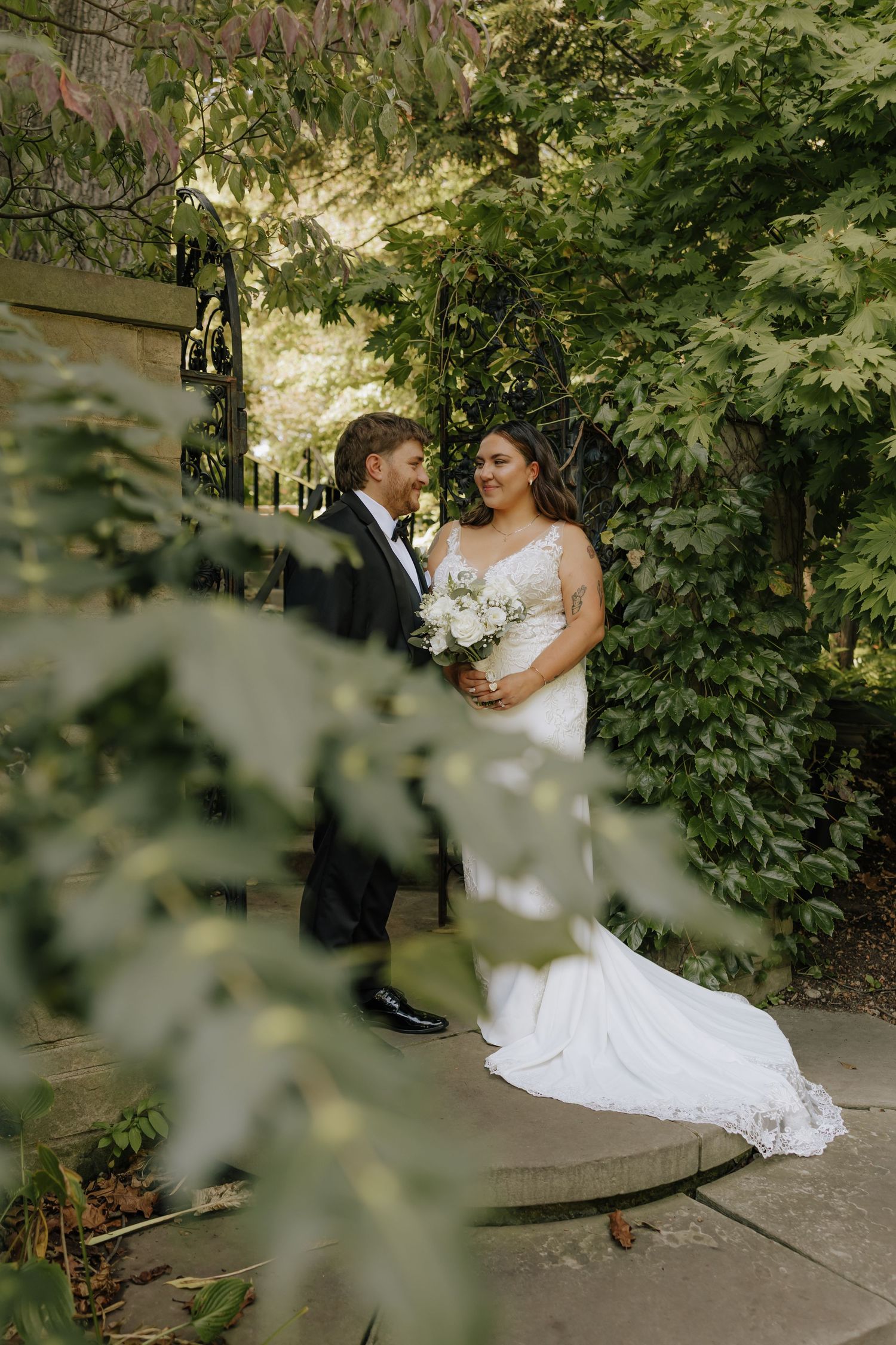 A romantic wedding couple poses together on a garden path framed by lush green tree