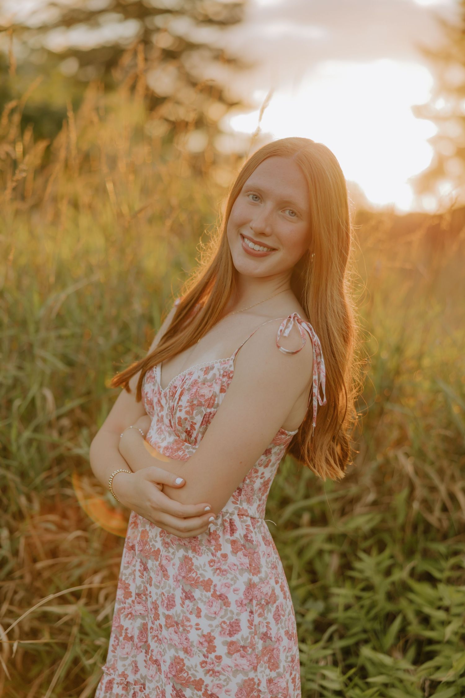 A young person in a floral dress poses in a field at sunset with warm golden lighting.