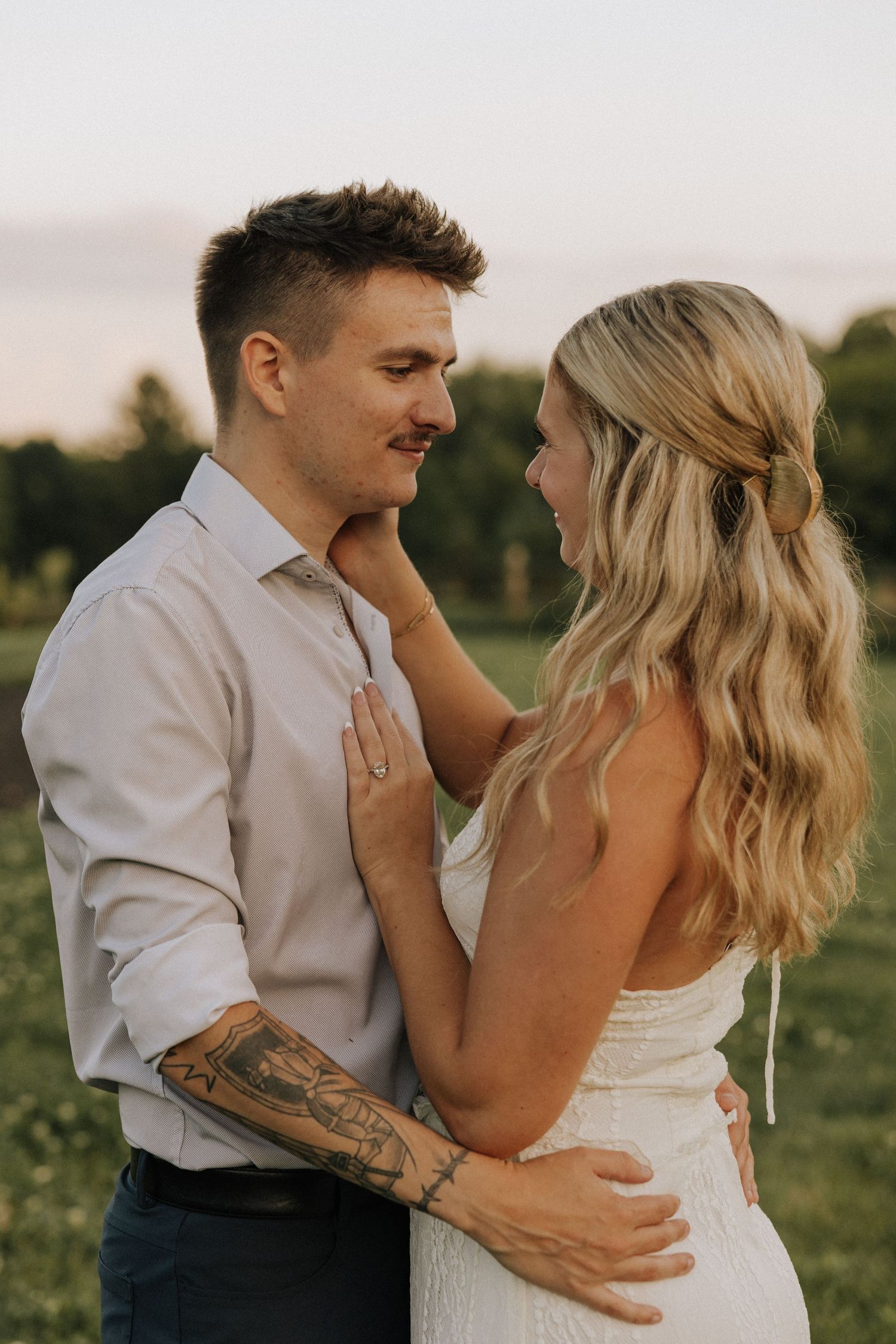 A couple shares a romantic moment outdoors at golden hour, the woman in white touching the mans face as he embraces her waist.