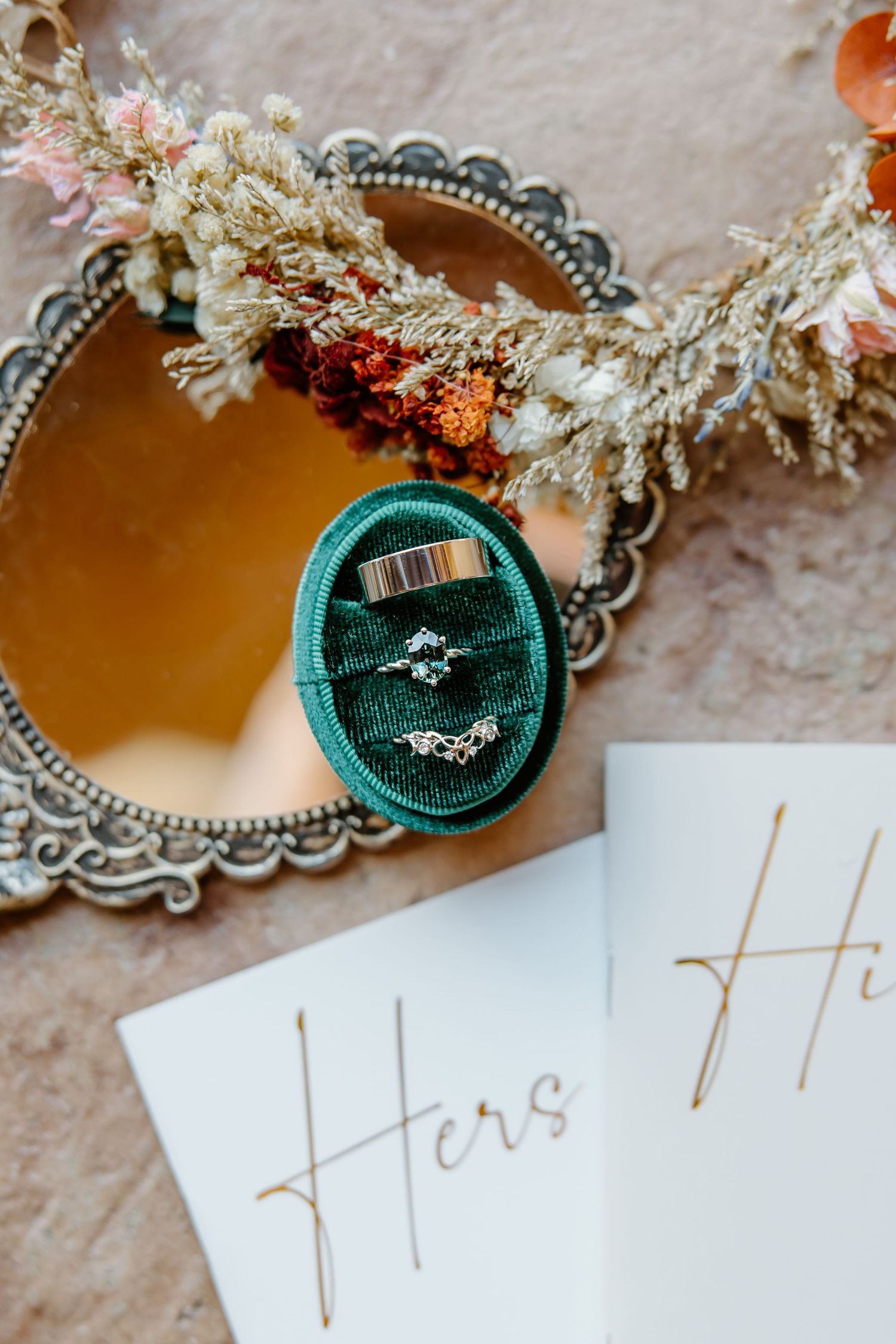 Teal velvet ring box surrounded by delicate dried flowers and calligraphy on white paper.