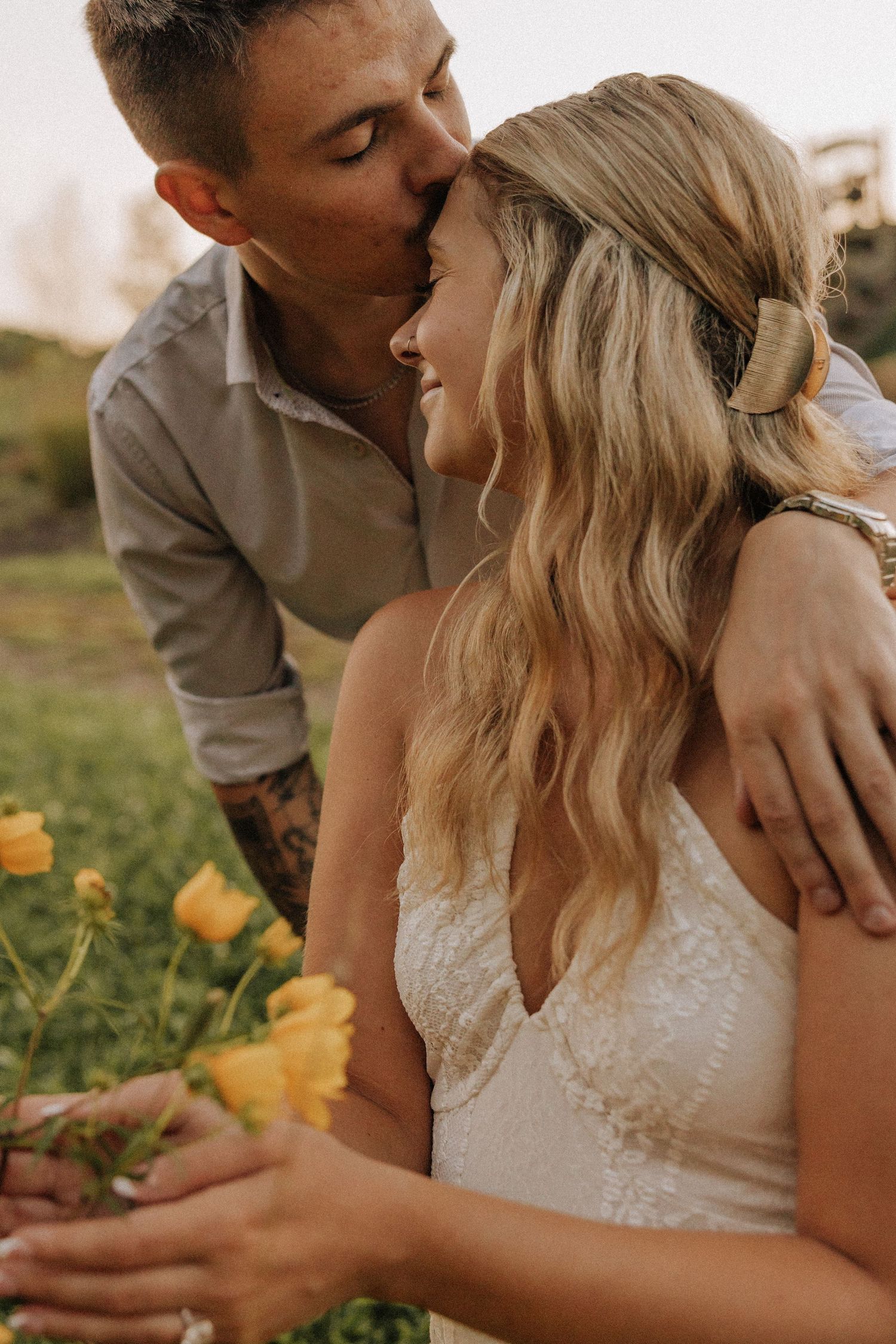 A groom with tattoos kisses his smiling blonde bride wearing a white lace dress among yellow flowers outdoors.