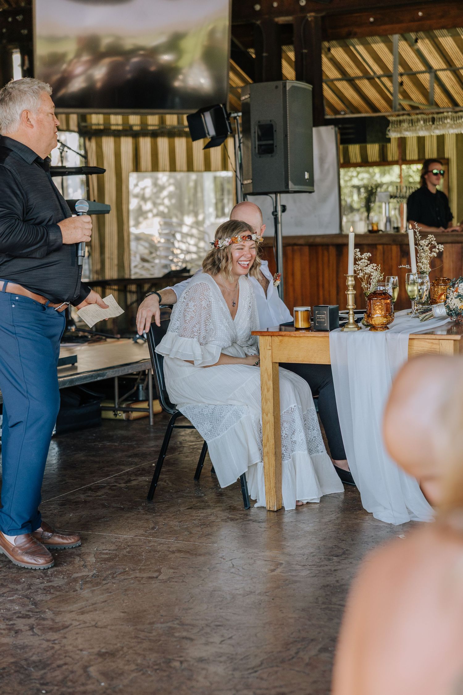 A wedding reception scene in a rustic covered venue with guests gathered around wooden tables.