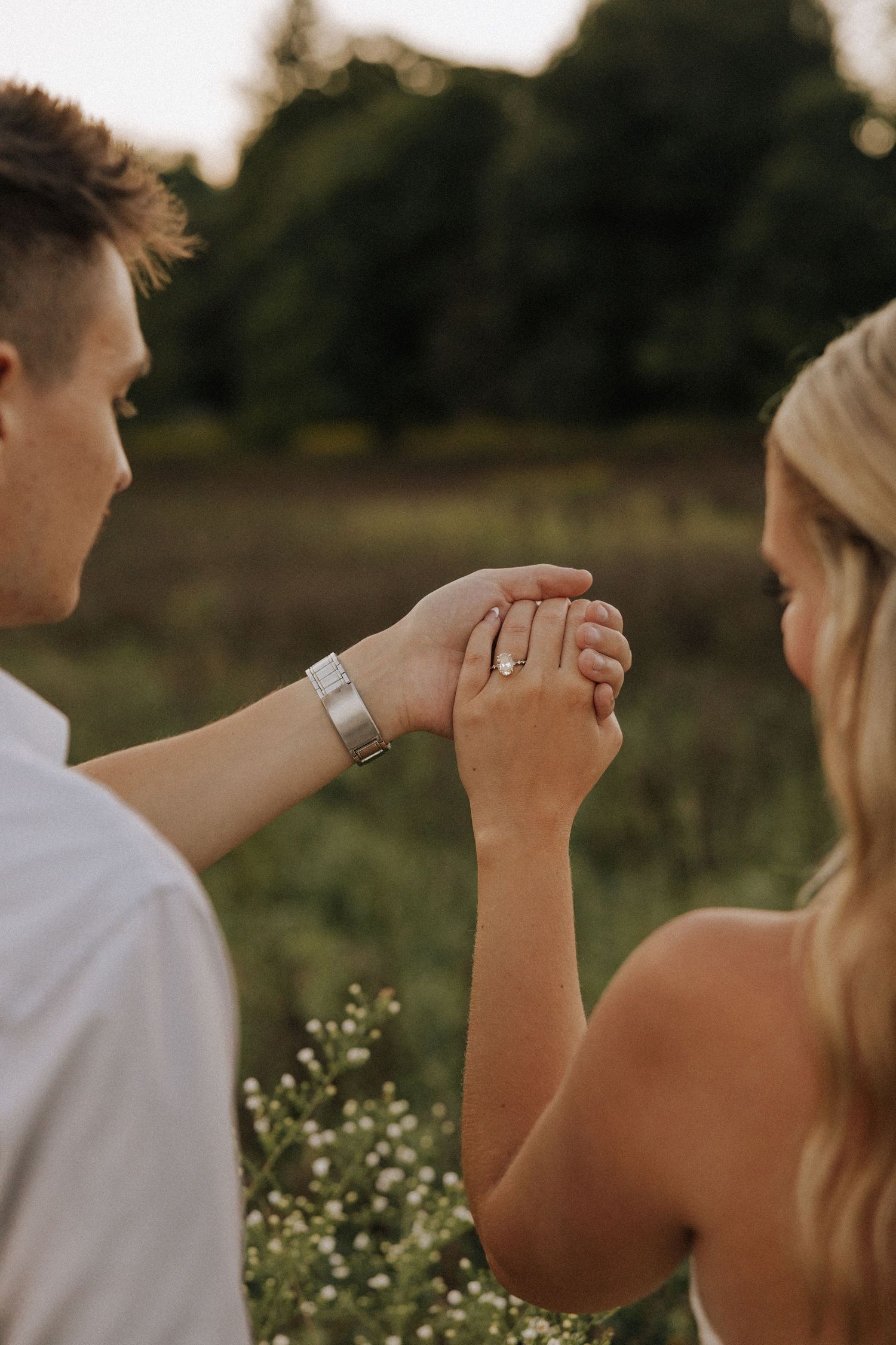 Couple holds hands showing diamond engagement ring and silver watch in outdoor garden setting.