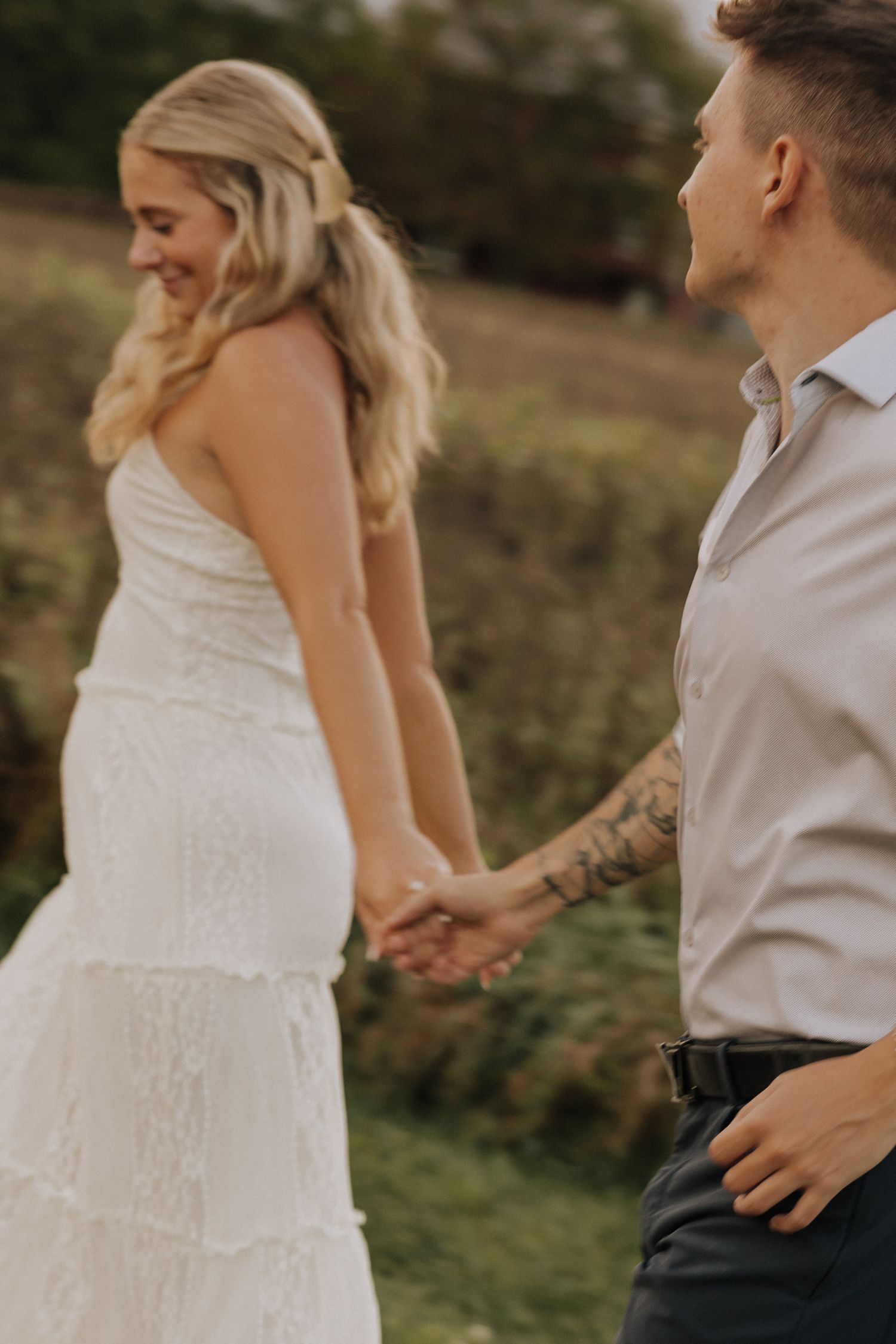 Bride in white lace gown holds hands with tattooed groom in white shirt walking through green field.
