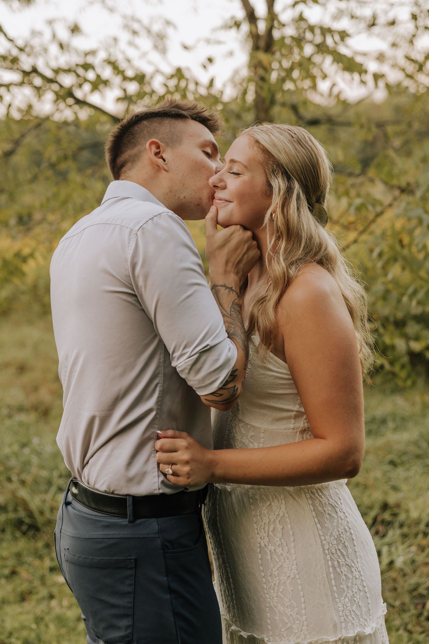 A couple shares a romantic kiss outdoors, with the man gently holding the woman's face in a lush green setting.