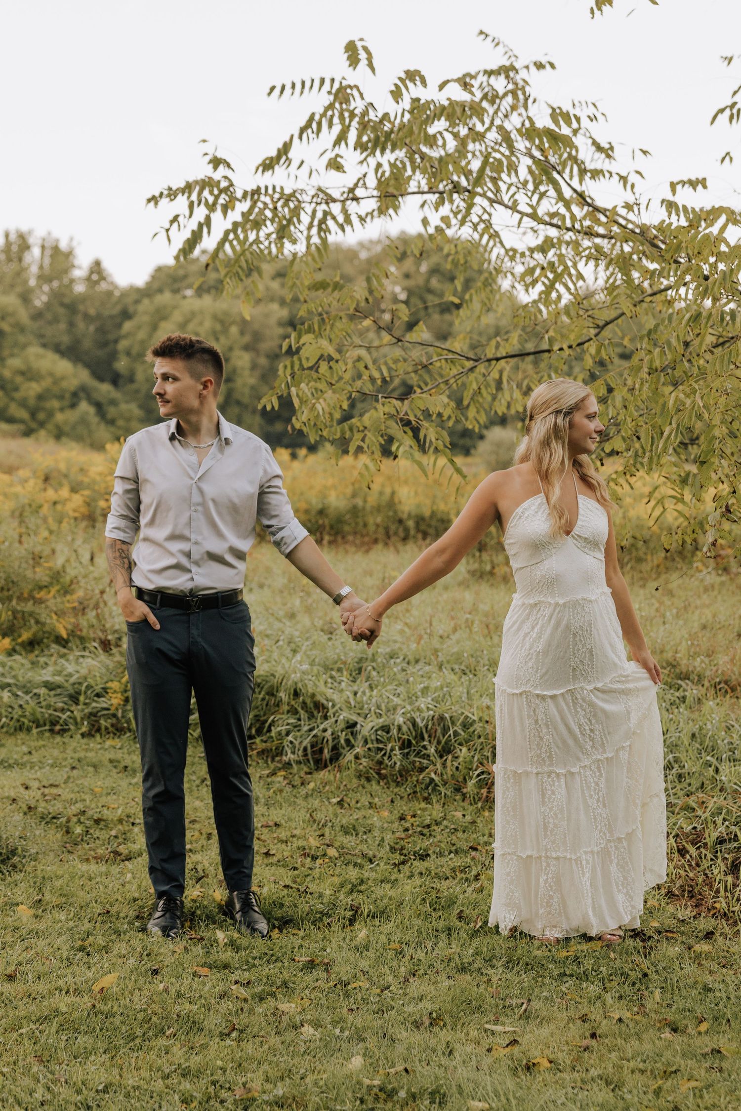 Couple holding hands in a field, woman in white lace dress, man in gray shirt and dark pants, outdoor engagement photo.