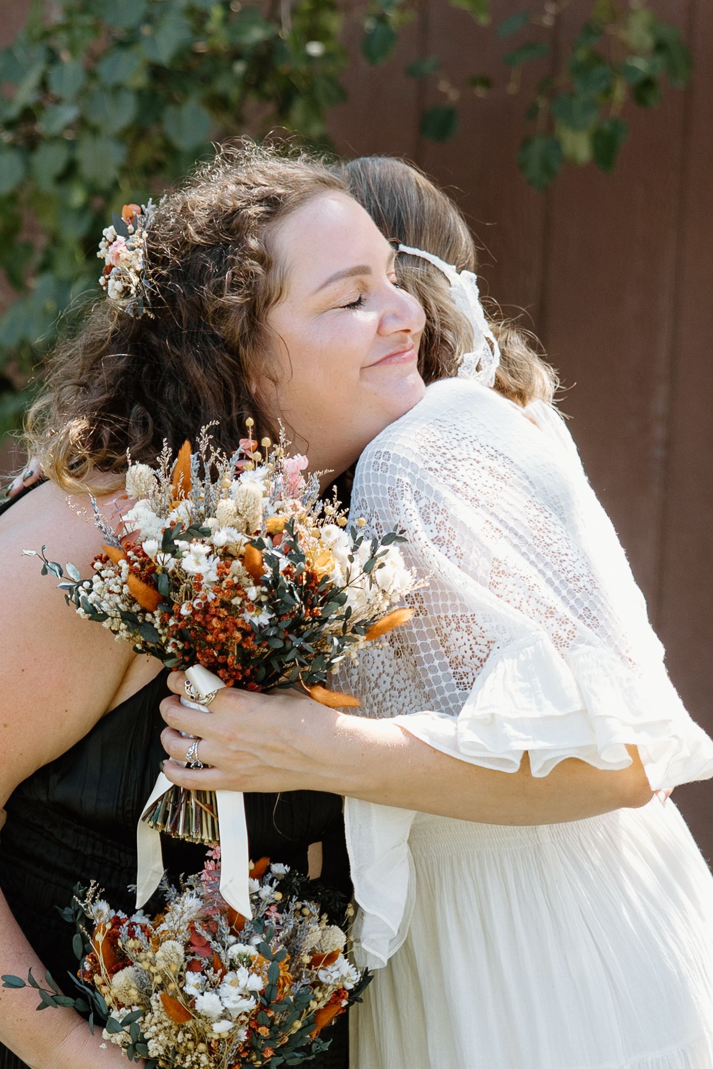 Two people in dresses share an emotional embrace while holding colorful dried flower bouquets during a wedding ceremony.