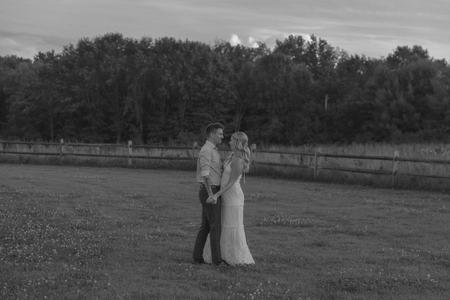 Black and white photo of a couple holding hands in a field, with trees and a fence in the background.