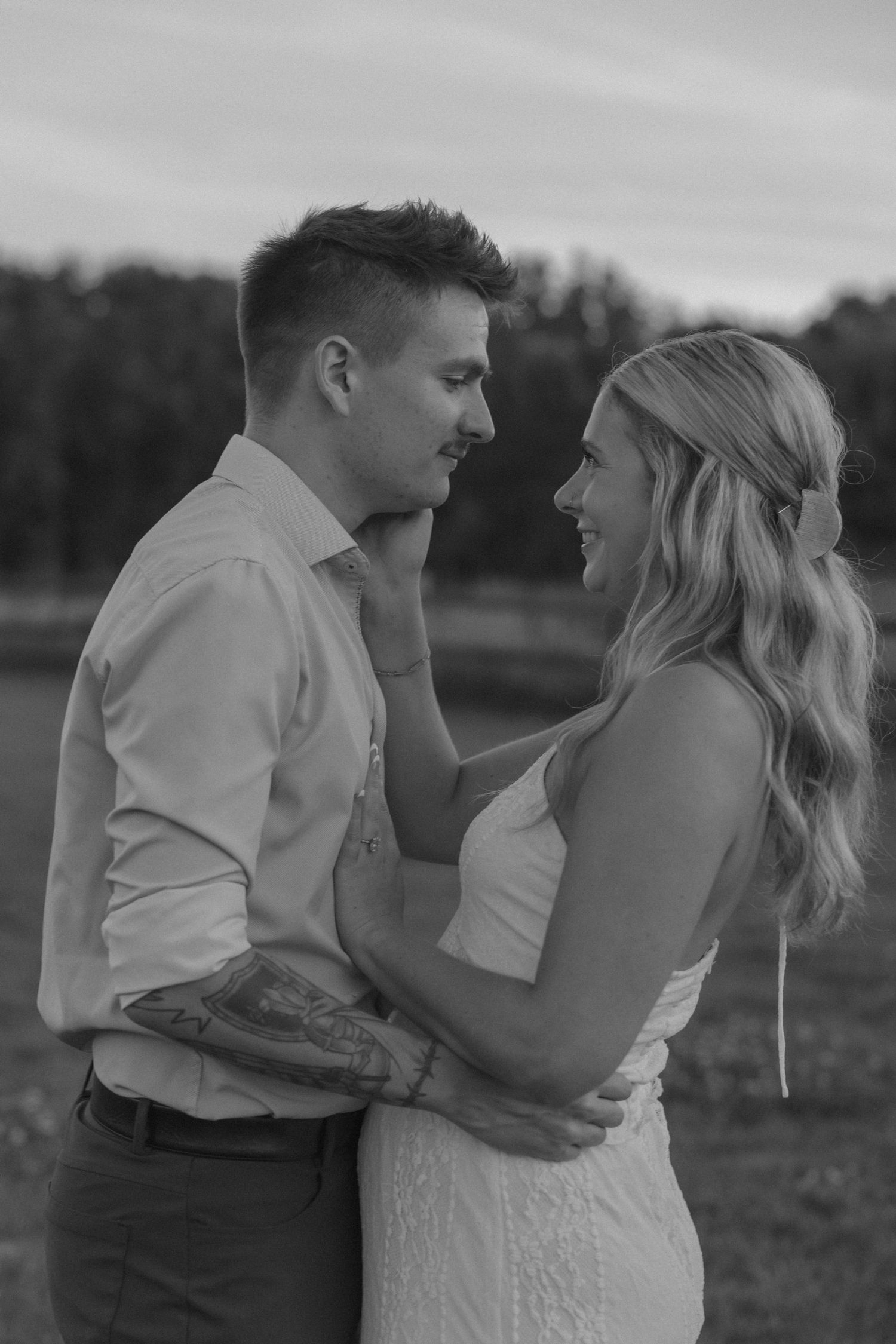 Black and white photo of smiling couple in wedding attire embracing outdoors at dusk.