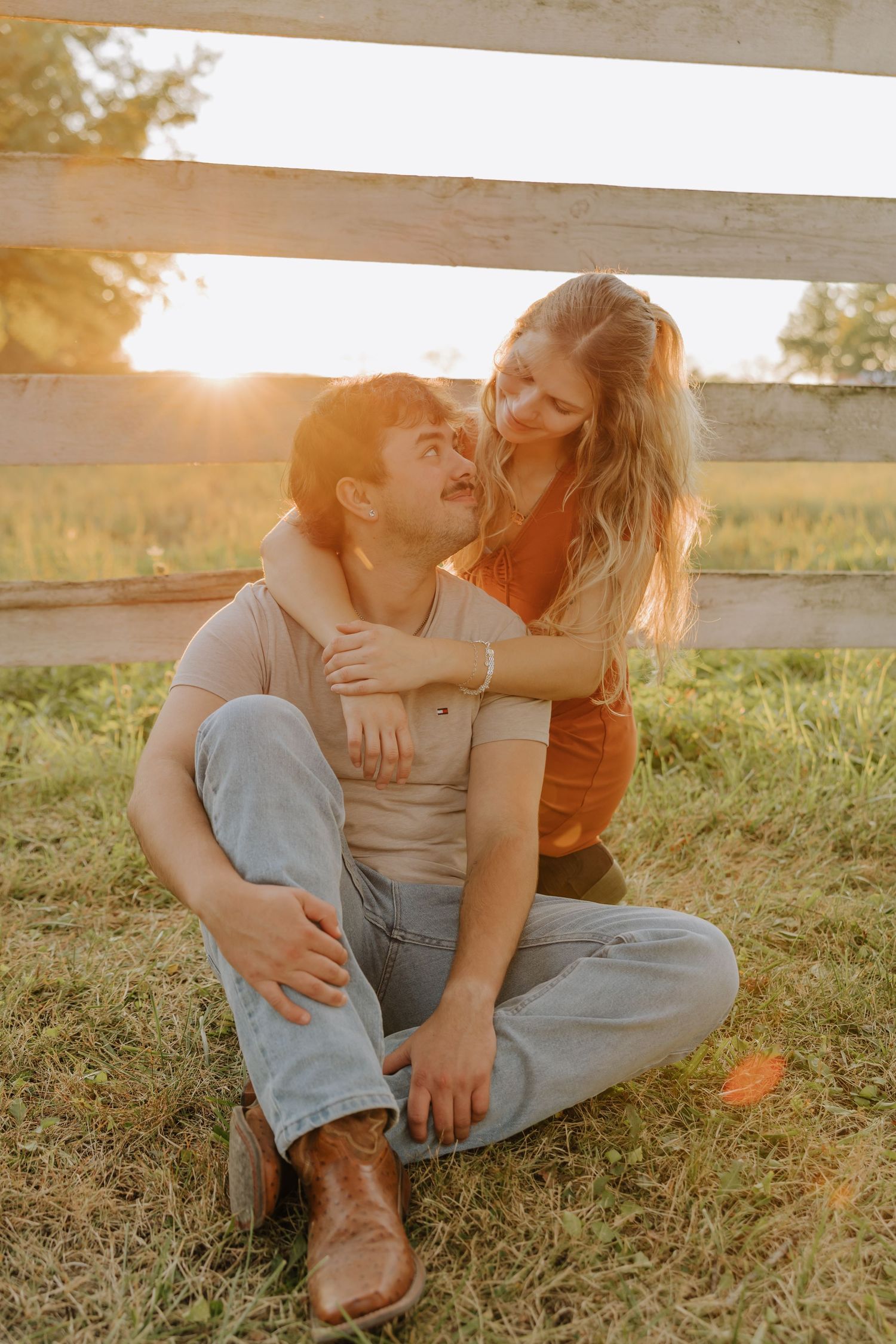 A couple sits together on grass during golden hour sunlight, sharing an intimate moment in a rural setting.