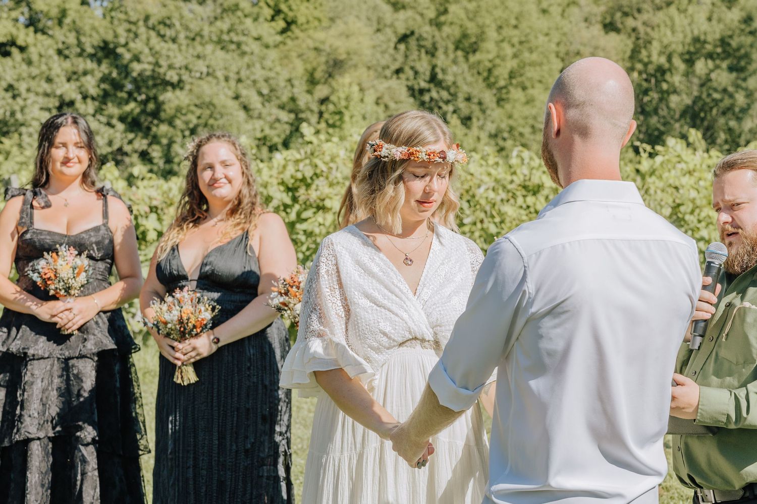 An outdoor wedding ceremony takes place in a sunlit garden setting with attendants in black and white attire.