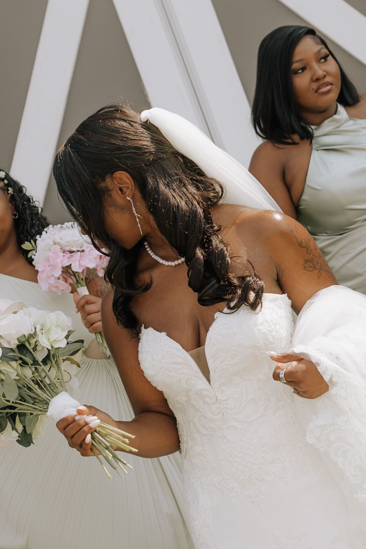 A bride in a white lace gown holds a pink bouquet surrounded by bridesmaids in sage green dresses at a wedding.