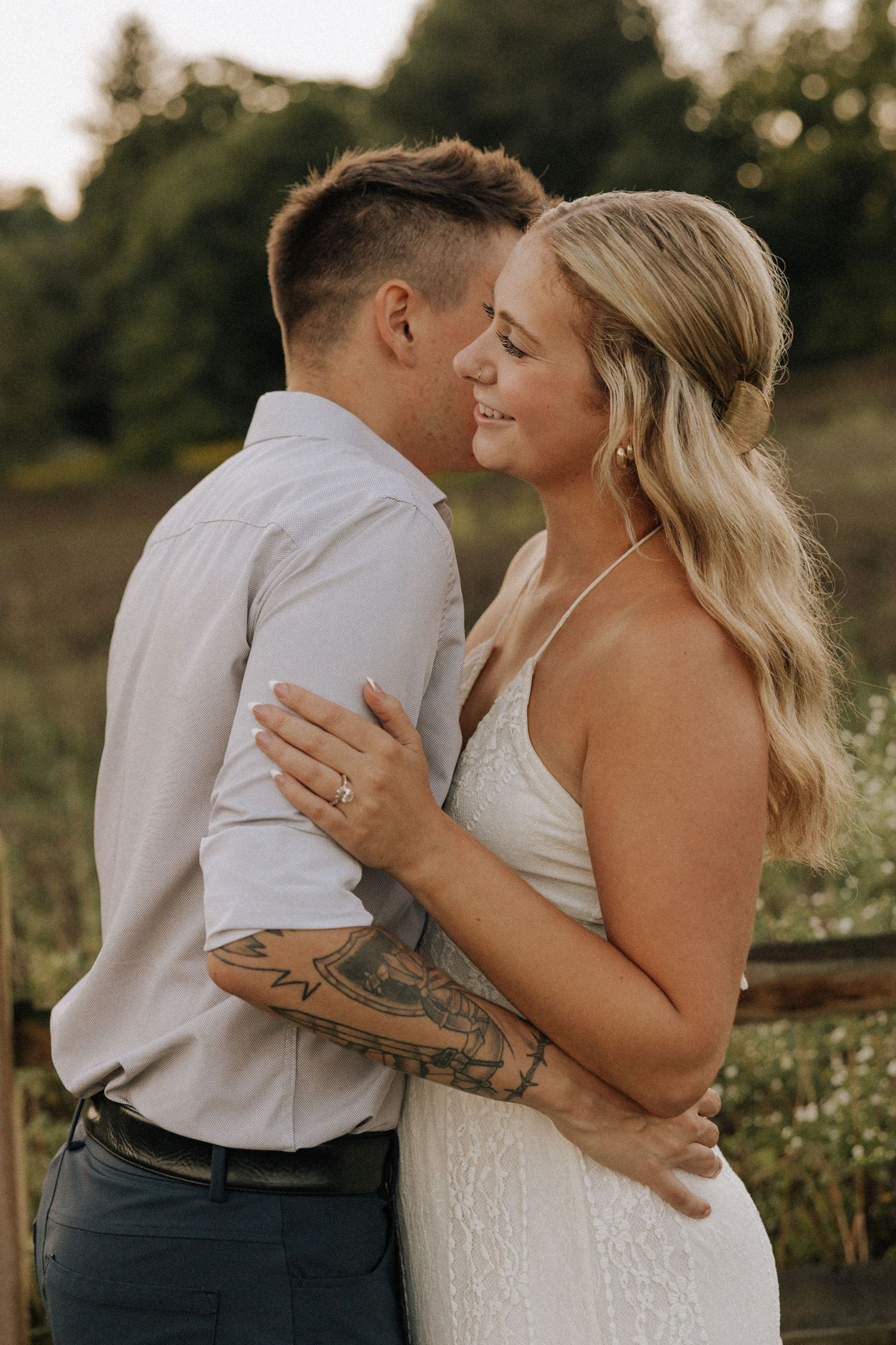 Couple in wedding attire embracing outdoors, bride in white halter gown with groom in white shirt, tattoos visible.
