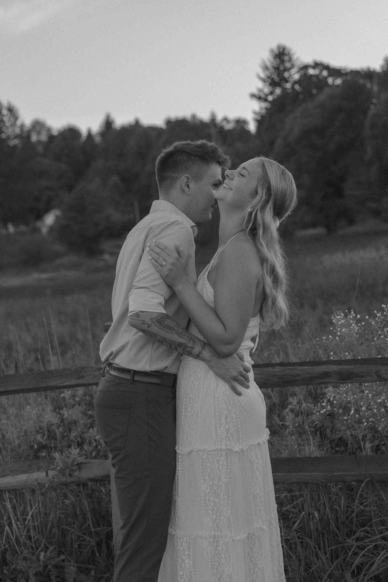 Black and white photo of couple almost kissing in a field at dusk, woman in white lace dress, man in button-up shirt.