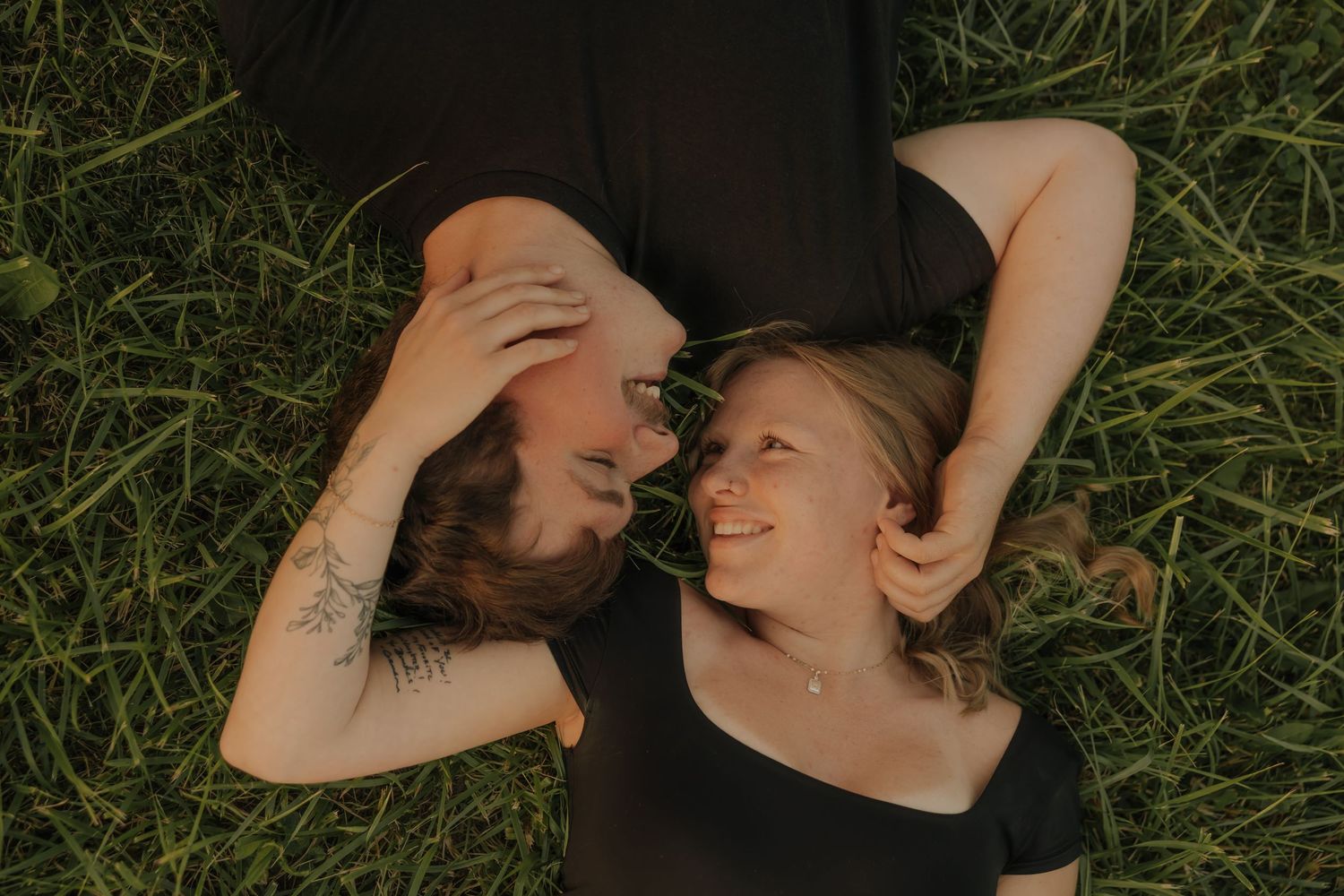 Two people lying on green grass, smiling at each other in a warm, intimate moment outdoors.