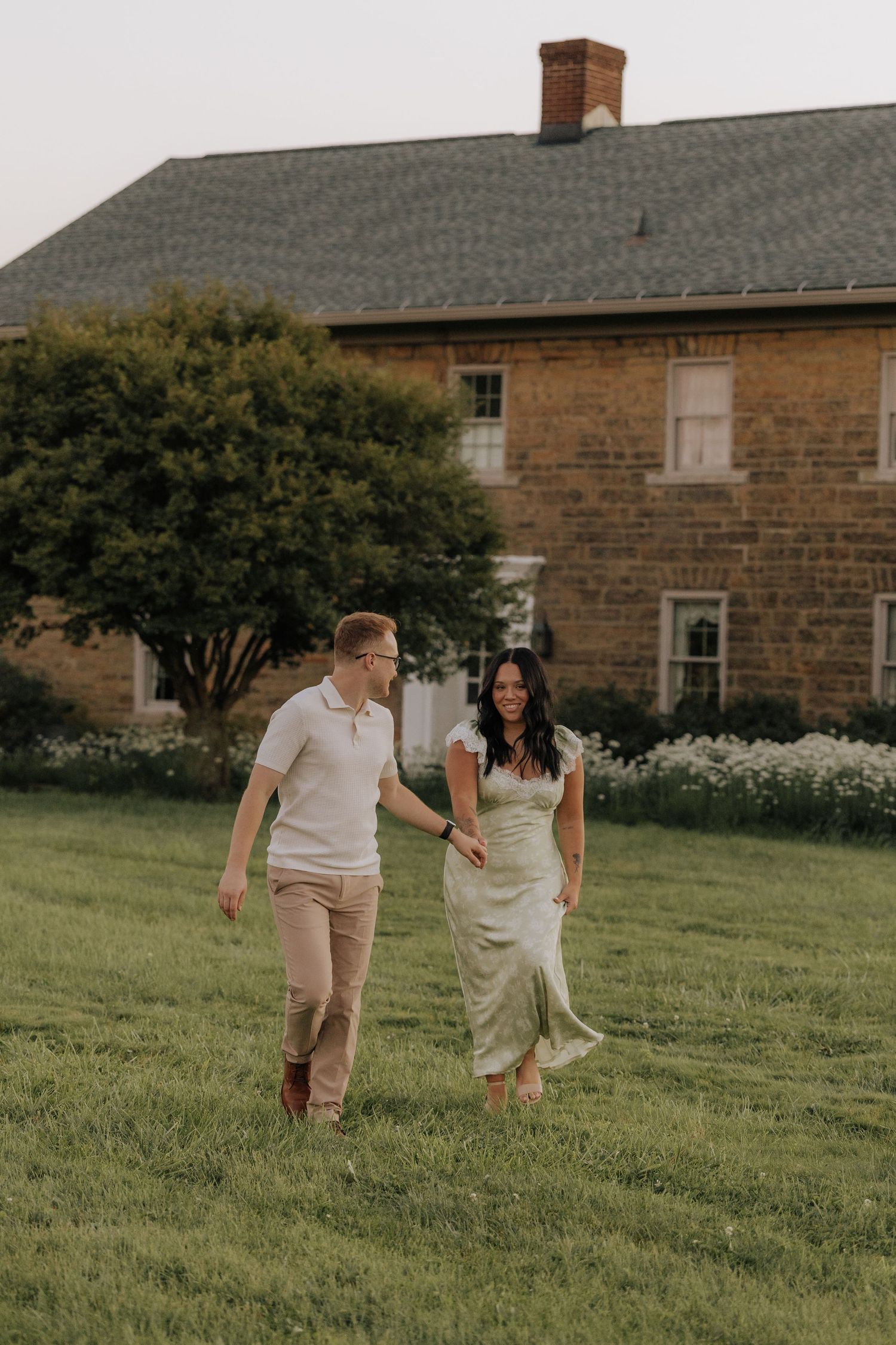 A couple walks hand-in-hand across a green lawn in front of a historic stone farmhouse at golden hour.