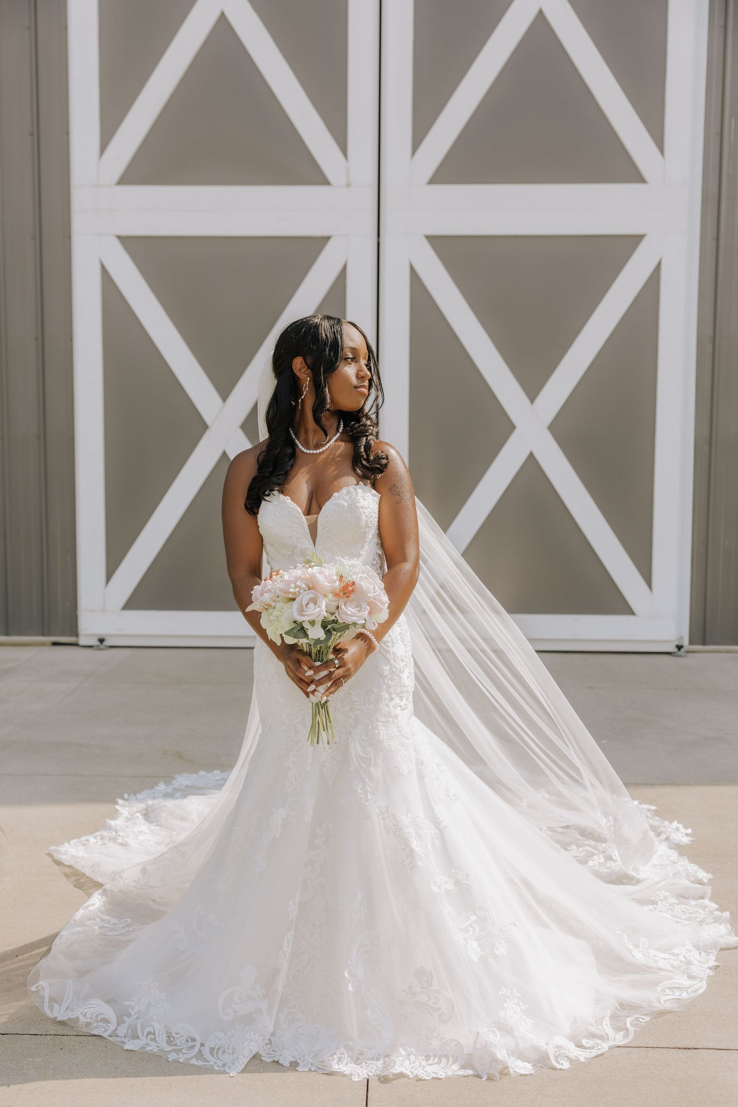 Elegant bride in a lace mermaid wedding gown holding a blush bouquet, posing before white barn doors.