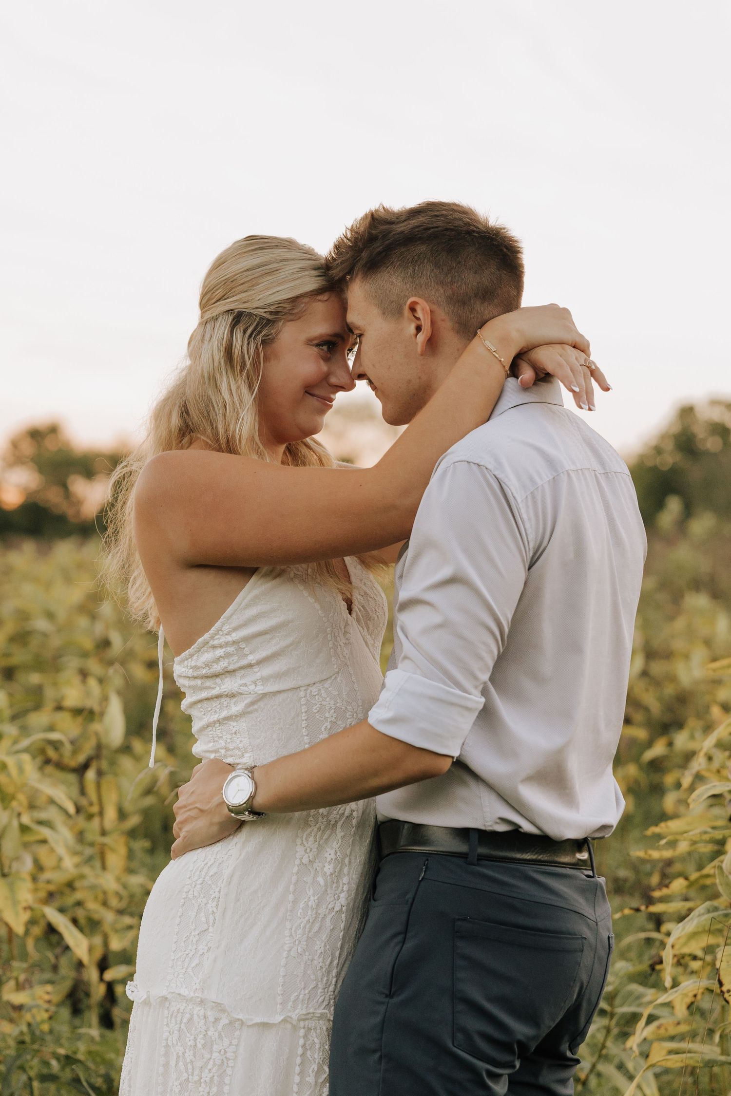 A couple in a sunflower field shares a romantic forehead touch, woman in white dress, man in white shirt.