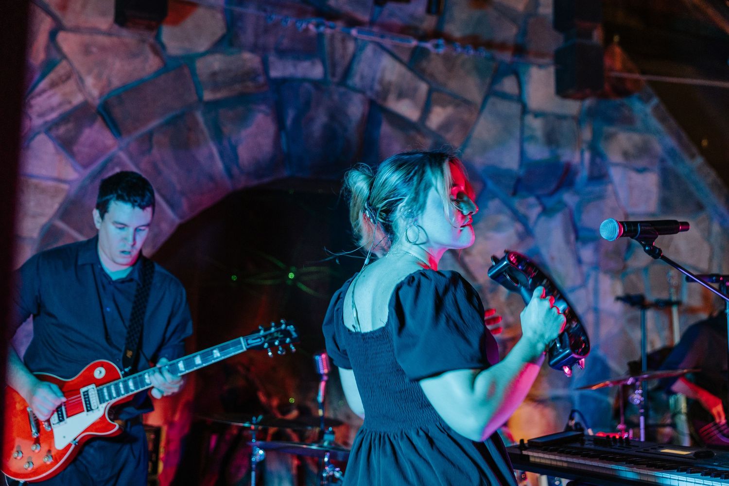 Musicians perform on stage with electric guitars in a dark venue lit by blue and red stage lighting.