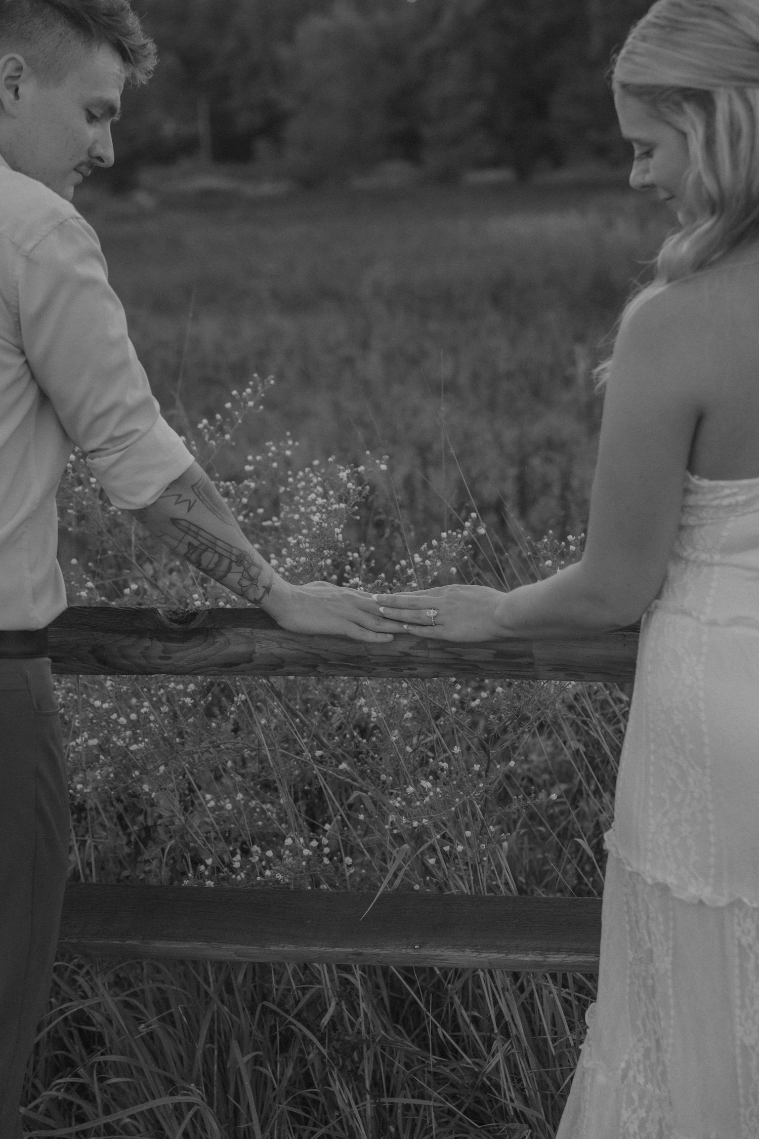 Black and white photo of bride and groom holding hands over rustic fence in field.