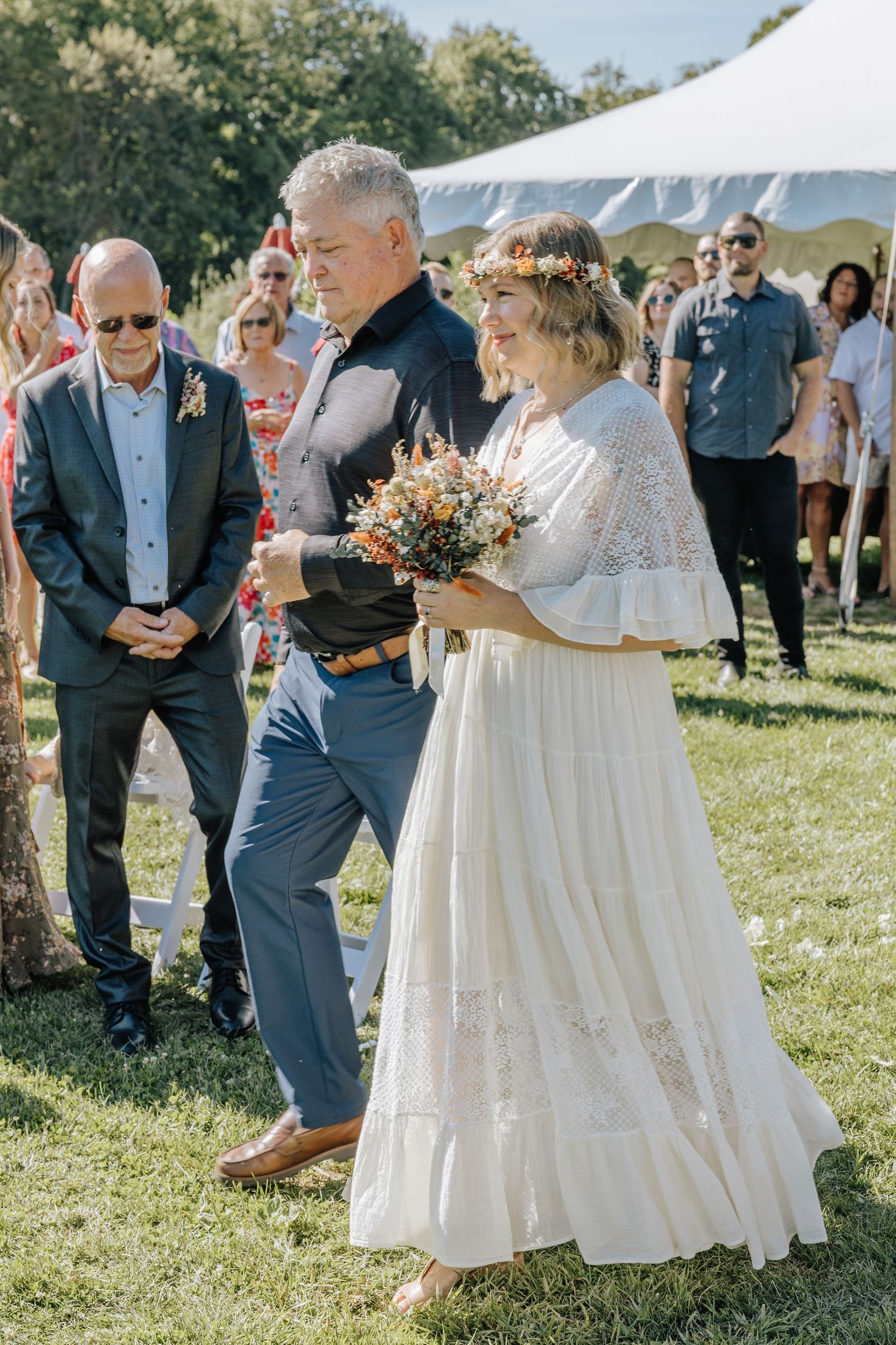 Wedding guests gather outdoors on a sunny day watching a bride walk down the aisle.