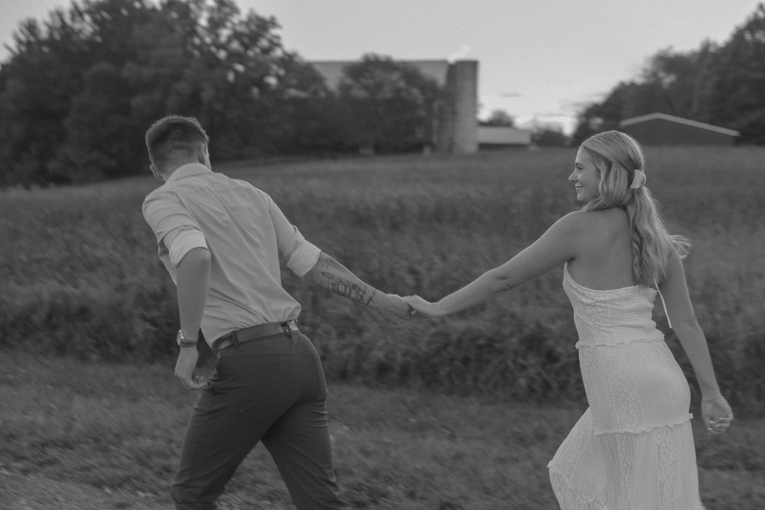 Black and white photo of a couple holding hands walking through a field near a barn at dusk.
