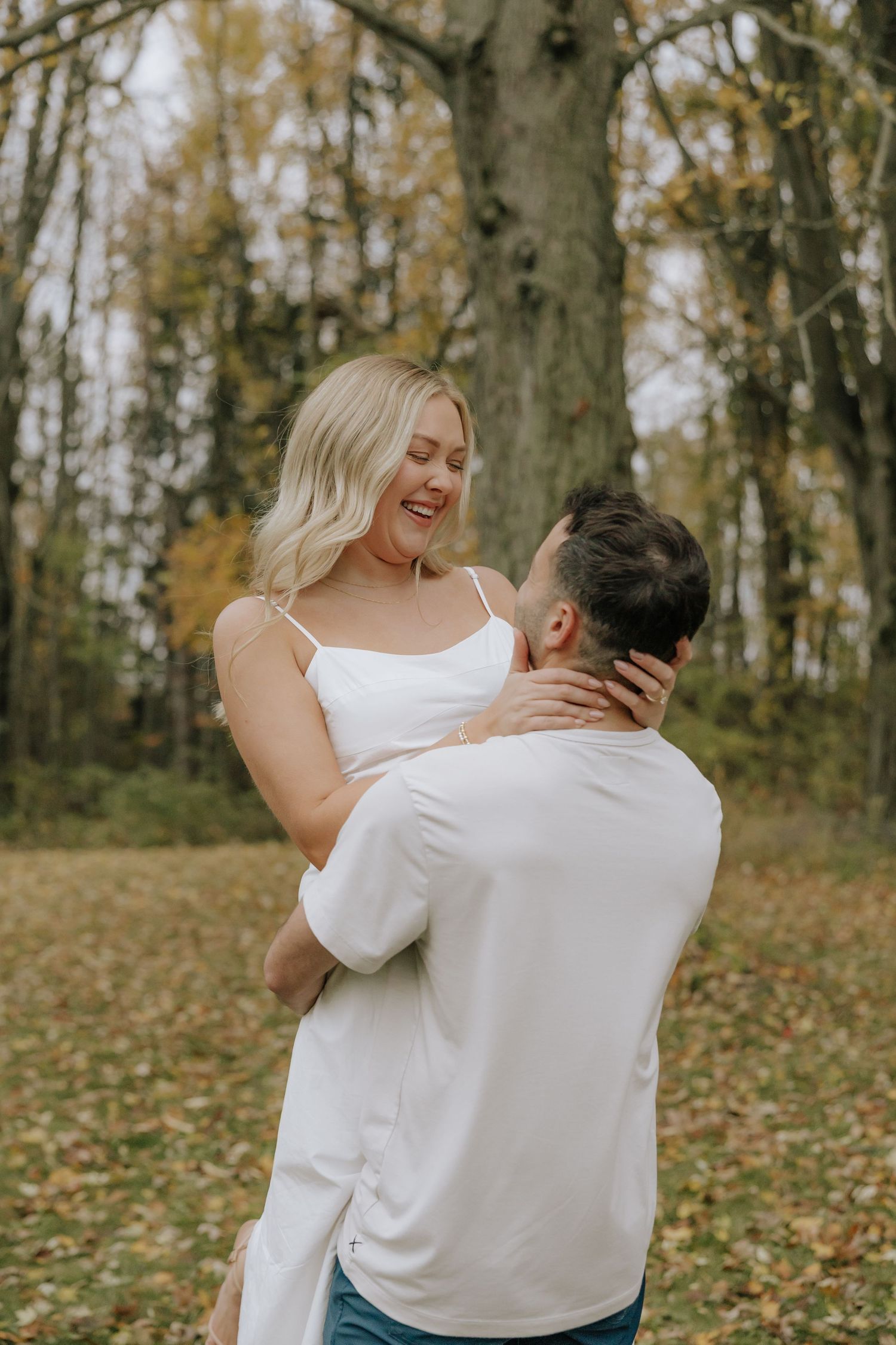 A couple shares a joyful embrace in an autumn forest setting with fallen leaves on the ground.