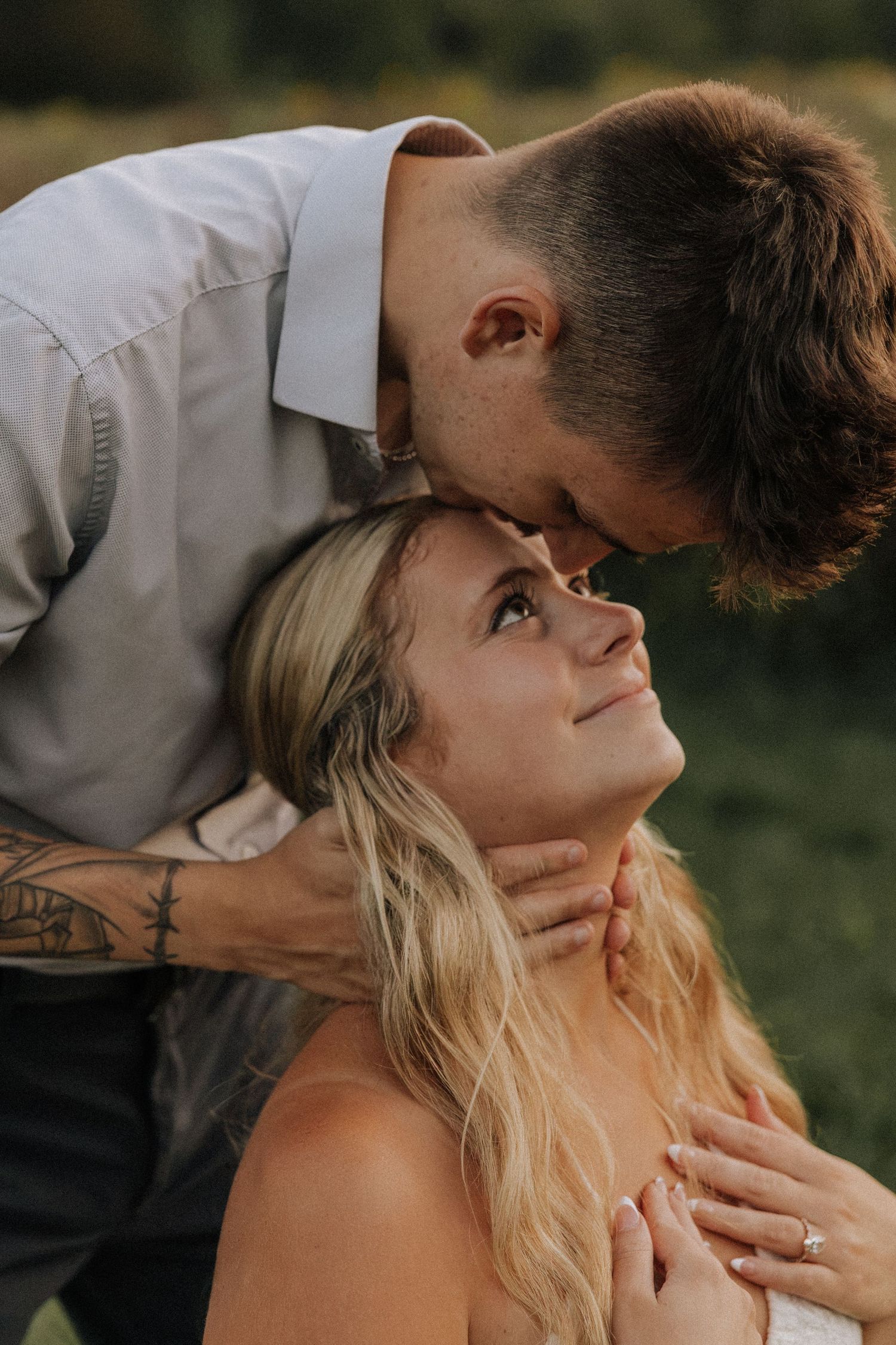 A groom with tattoos tenderly kisses his smiling blonde bride on the forehead during a romantic outdoor wedding.