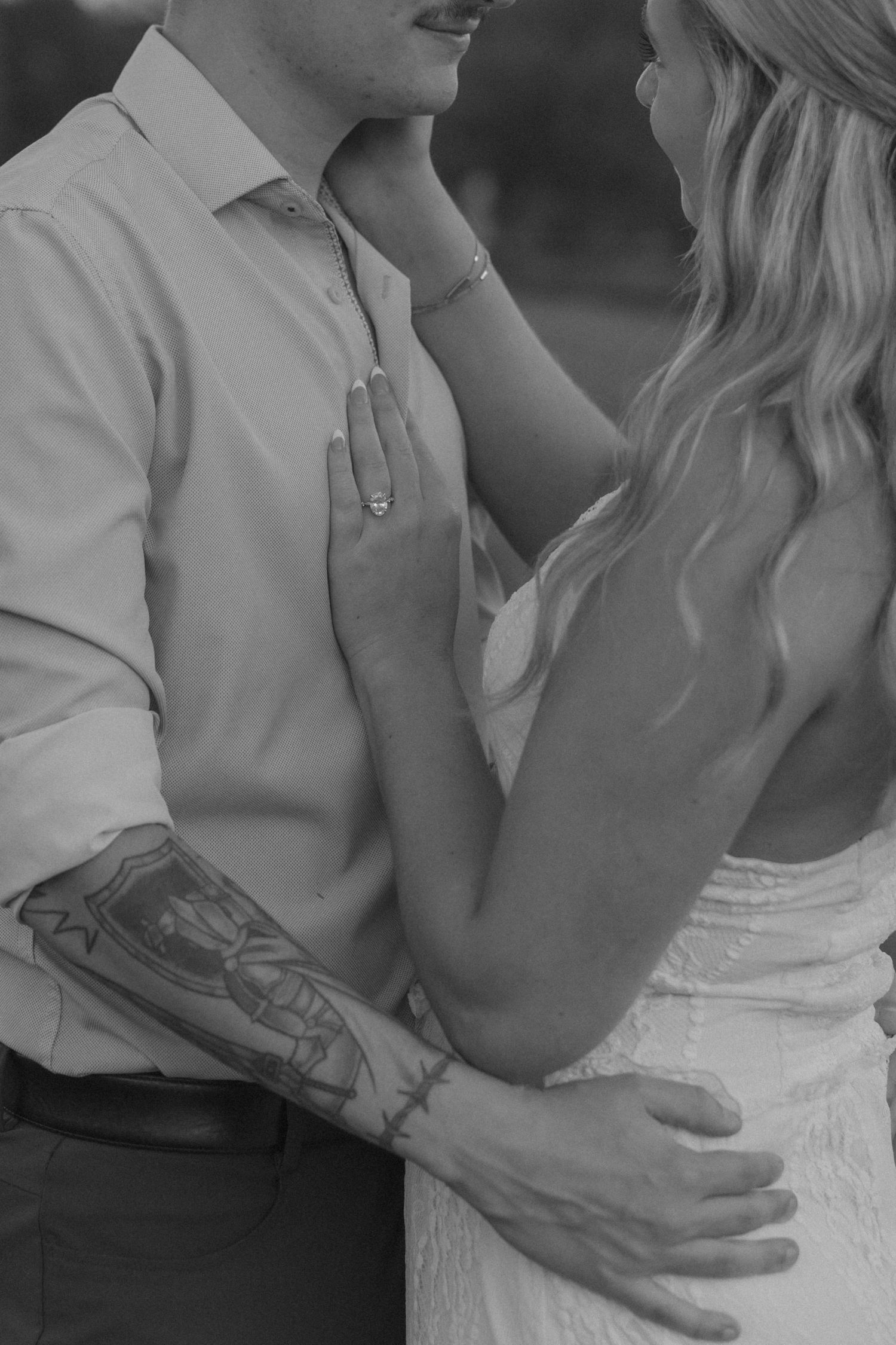 Black and white wedding photo of bride and groom embracing, showing tattooed arm, lace dress, and diamond engagement ring.