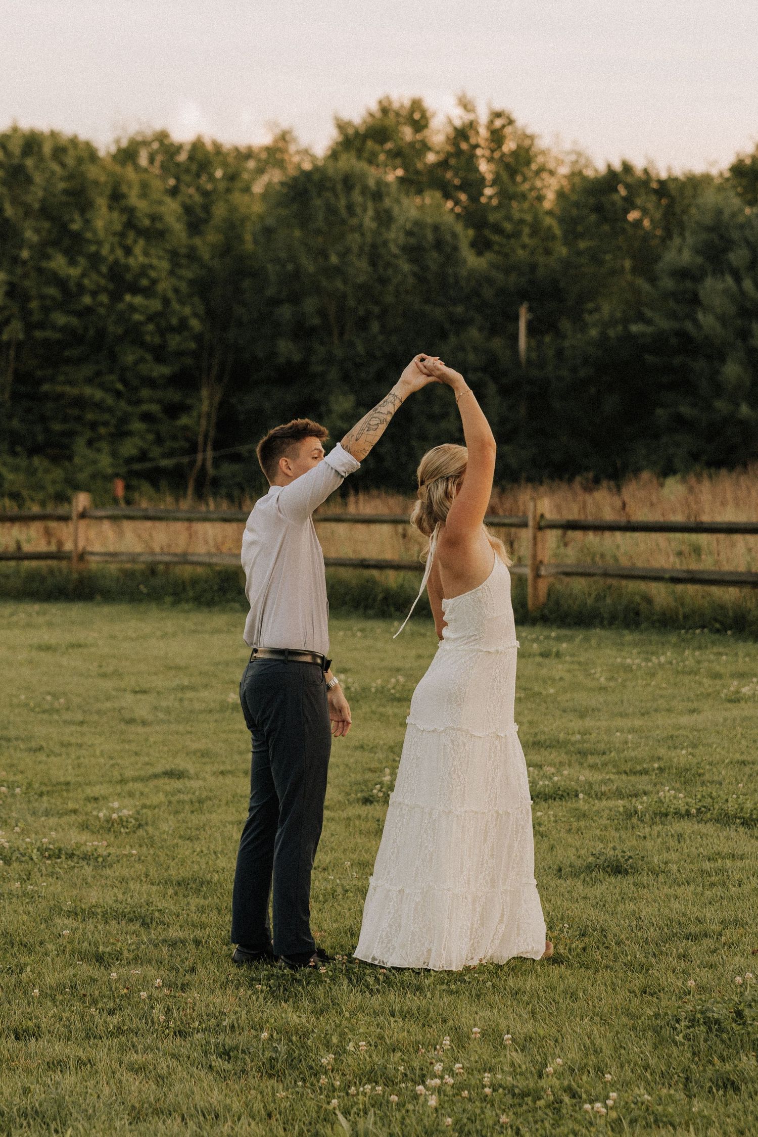 Bride and groom dancing outdoors at golden hour on green grass with wooden fence and trees in background.