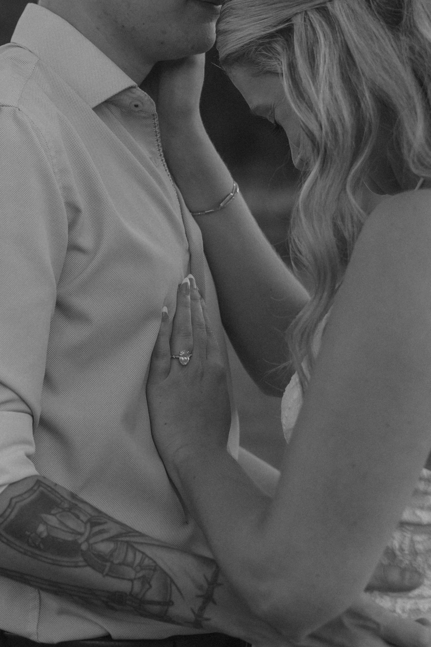 Black and white photo of an engaged couple embracing, showcasing a diamond ring and tattooed arm.