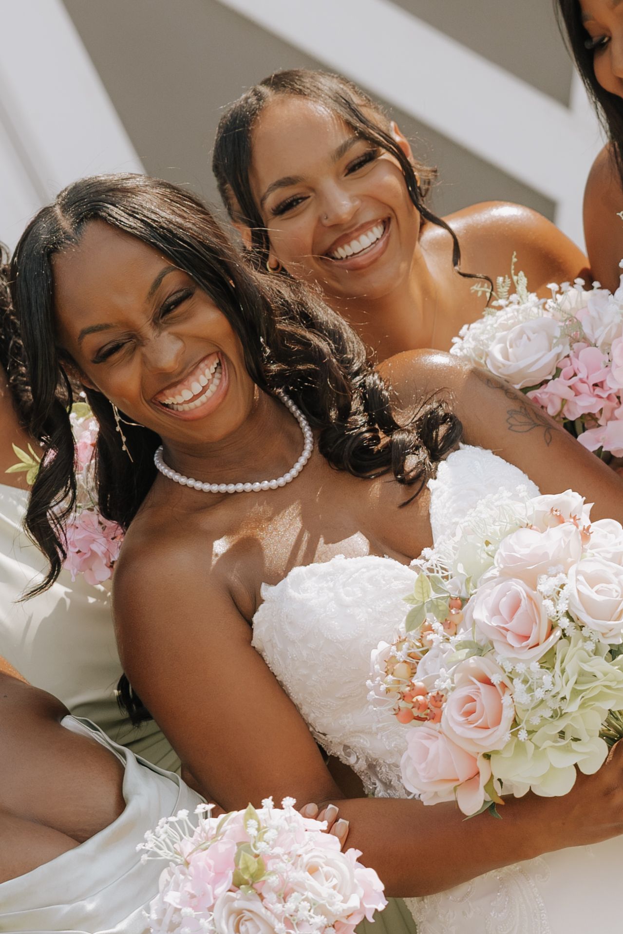 Two joyful brides smile and laugh together holding blush pink rose bouquets on a sunny wedding day.