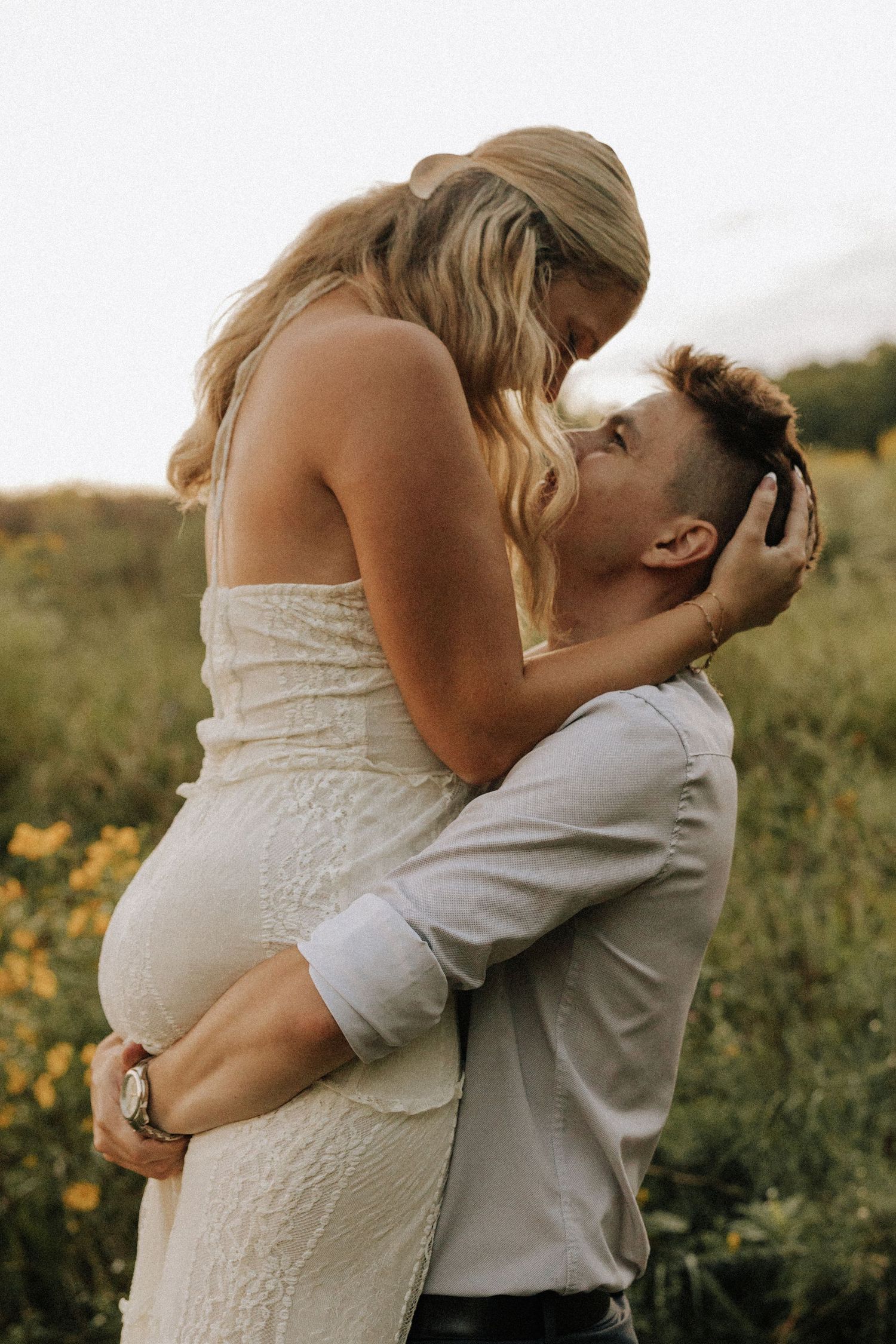 A man lifts a woman in a white dress as they share a romantic moment in a sunny wildflower field.