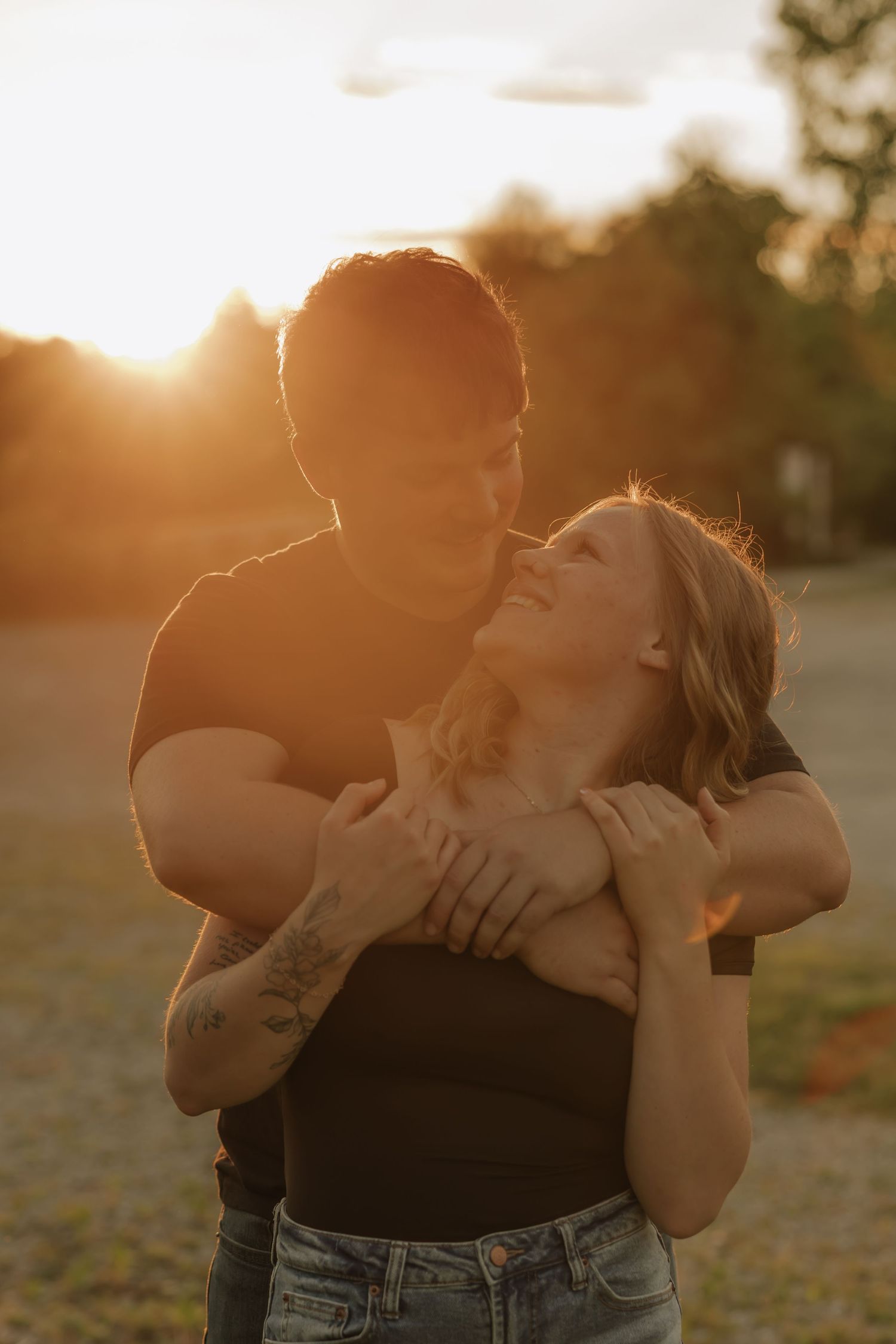A couple embraces at golden hour sunset, the man hugging the woman from behind as she laughs joyfully outdoors.