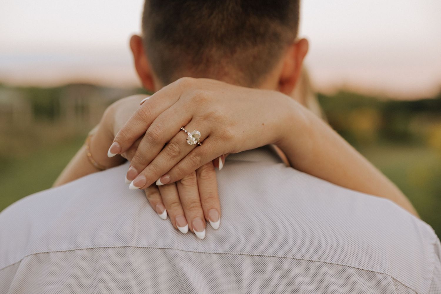 Close-up of a woman's hand with an engagement ring resting on a man's shoulder at sunset.